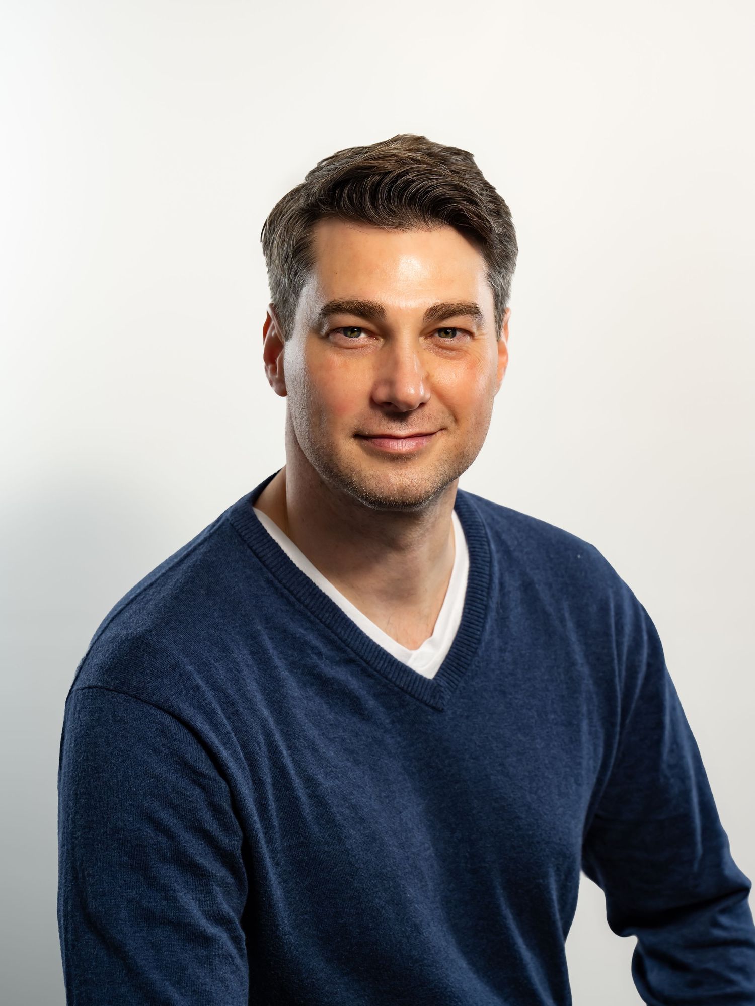 Professional headshot of a smiling man in a navy blue v-neck sweater against a light gray background.