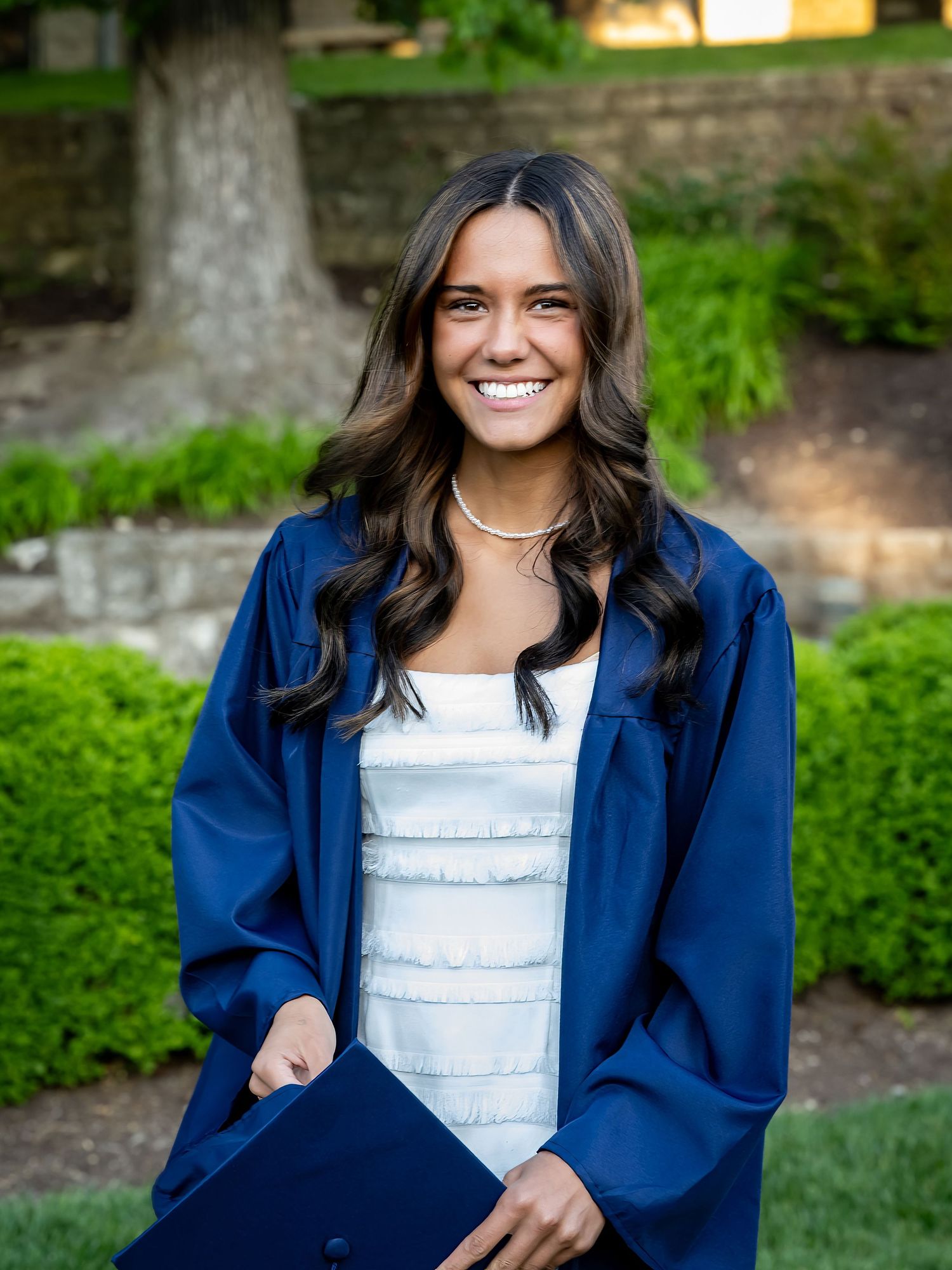 Graduate in navy blue regalia poses on stone steps with white dress underneath.