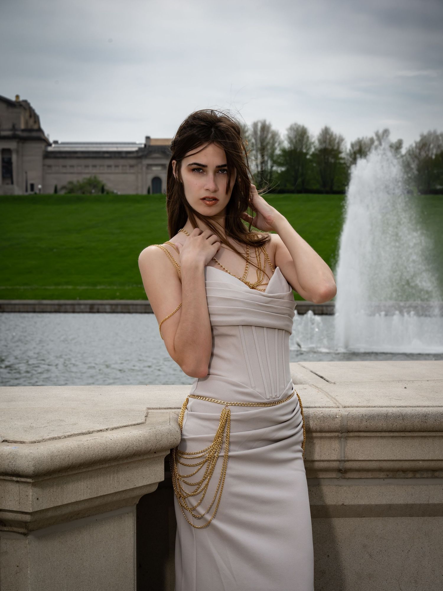 Model in beige dress stands elegantly by stone railing with fountain and green landscape in background.