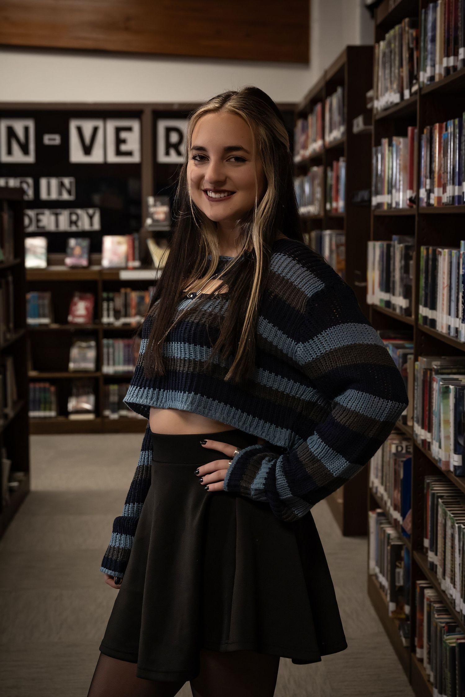 Person in dark sweater poses playfully between library bookshelves.
