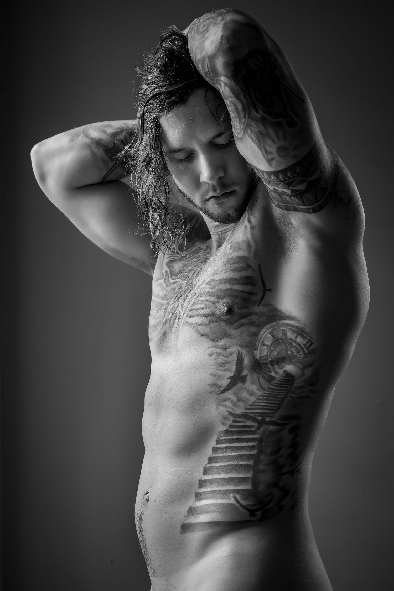 Black and white photo of a tattooed muscular man with long hair posing shirtless in a studio setting.