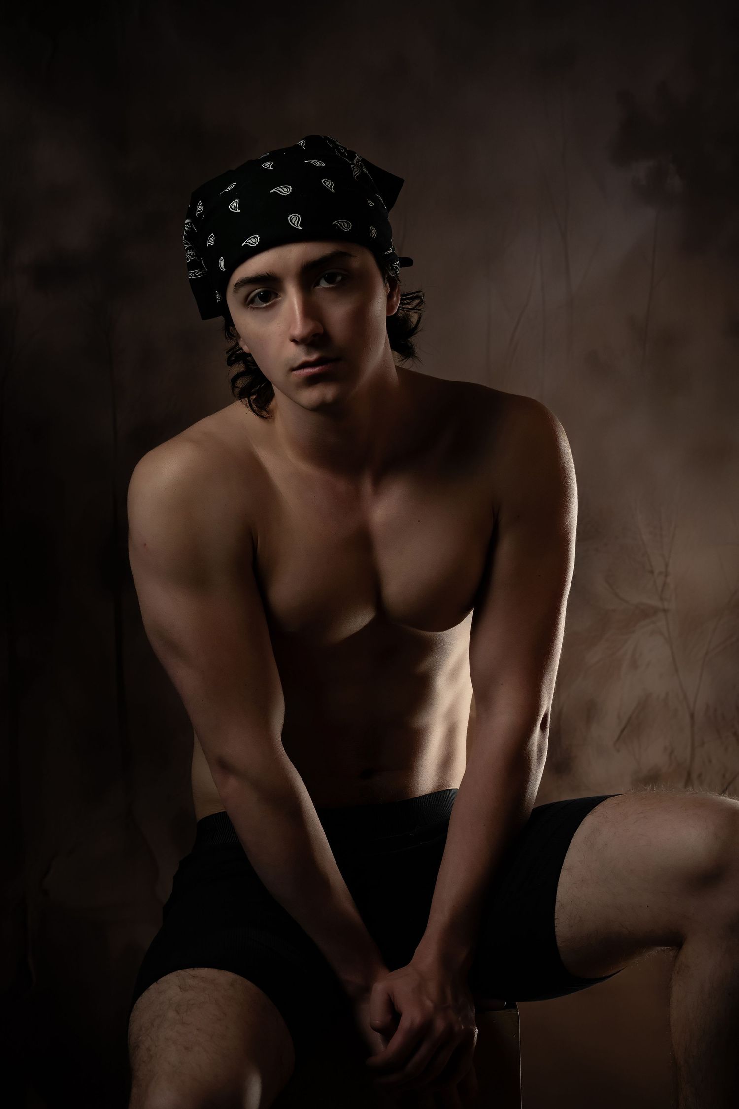 Artistic portrait of a figure wearing a patterned bandana and black shorts in moody lighting against dark background.