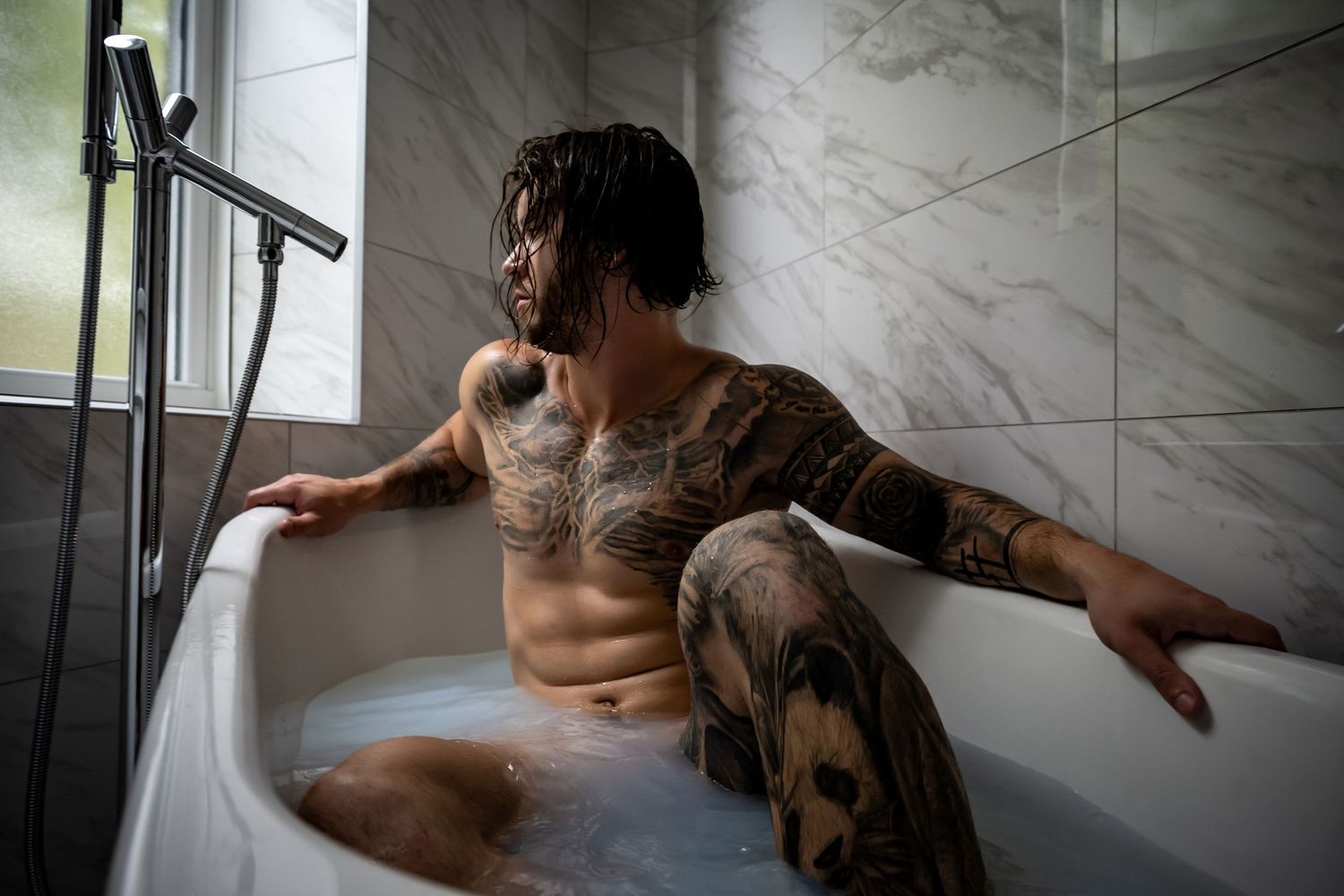 A tattooed person relaxes in a white bathtub near a window with natural light streaming in.