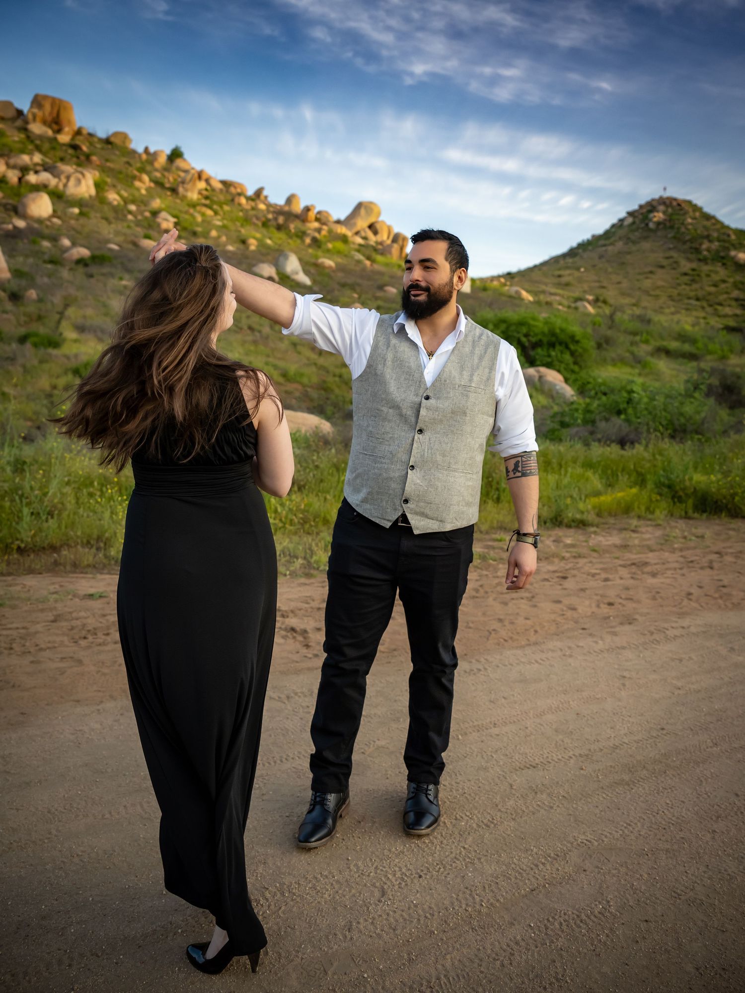 A couple in formal attire walking along a scenic mountain trail at sunset.
