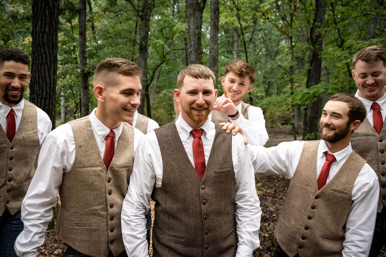 Groomsmen in brown vests and red ties pose together outdoors among trees.