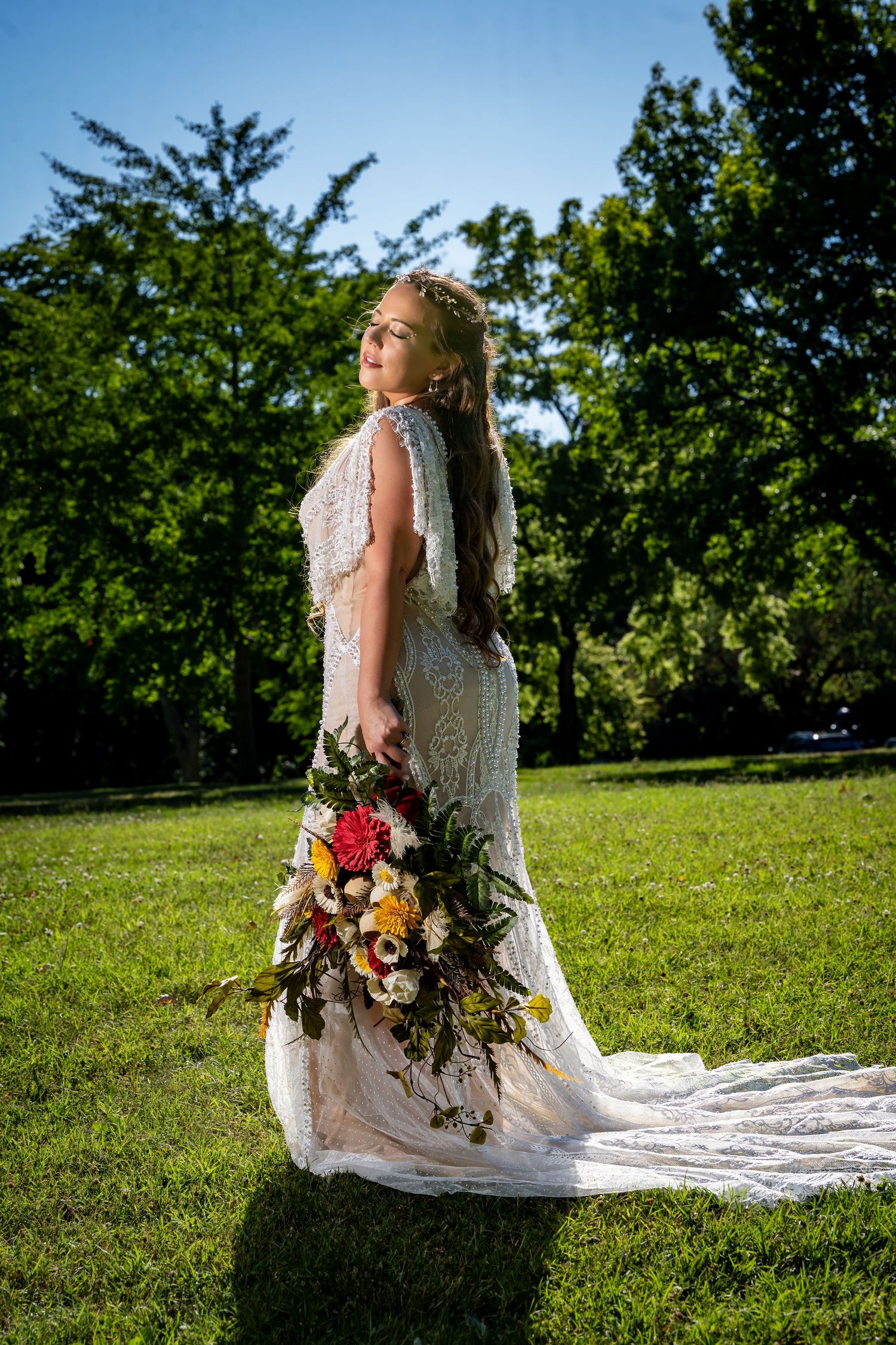 Bridal portrait in sunlit garden with flowing lace dress and colorful bouquet.