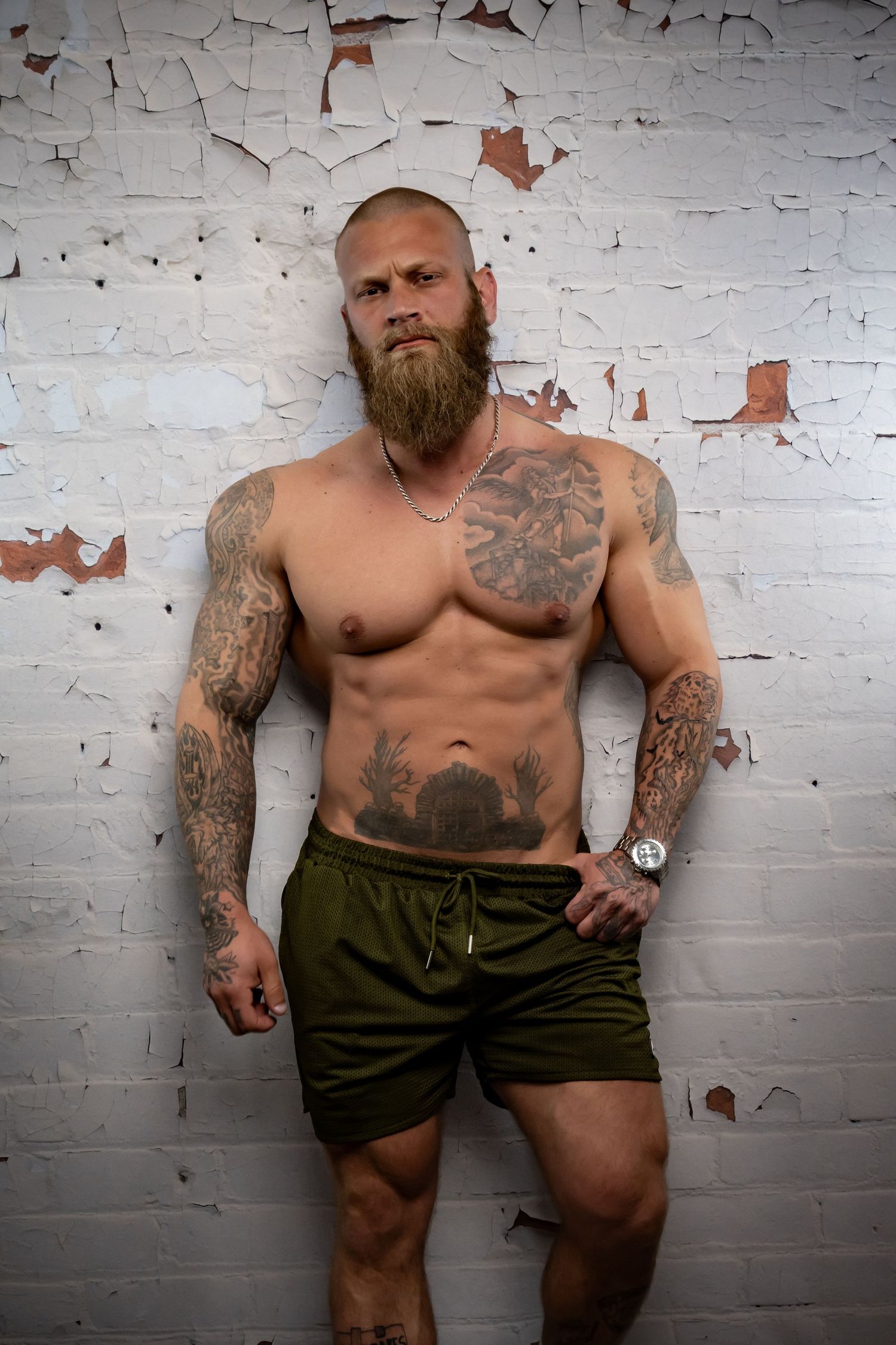 Muscular tattooed man with beard poses shirtless in green shorts against a rustic white brick wall.