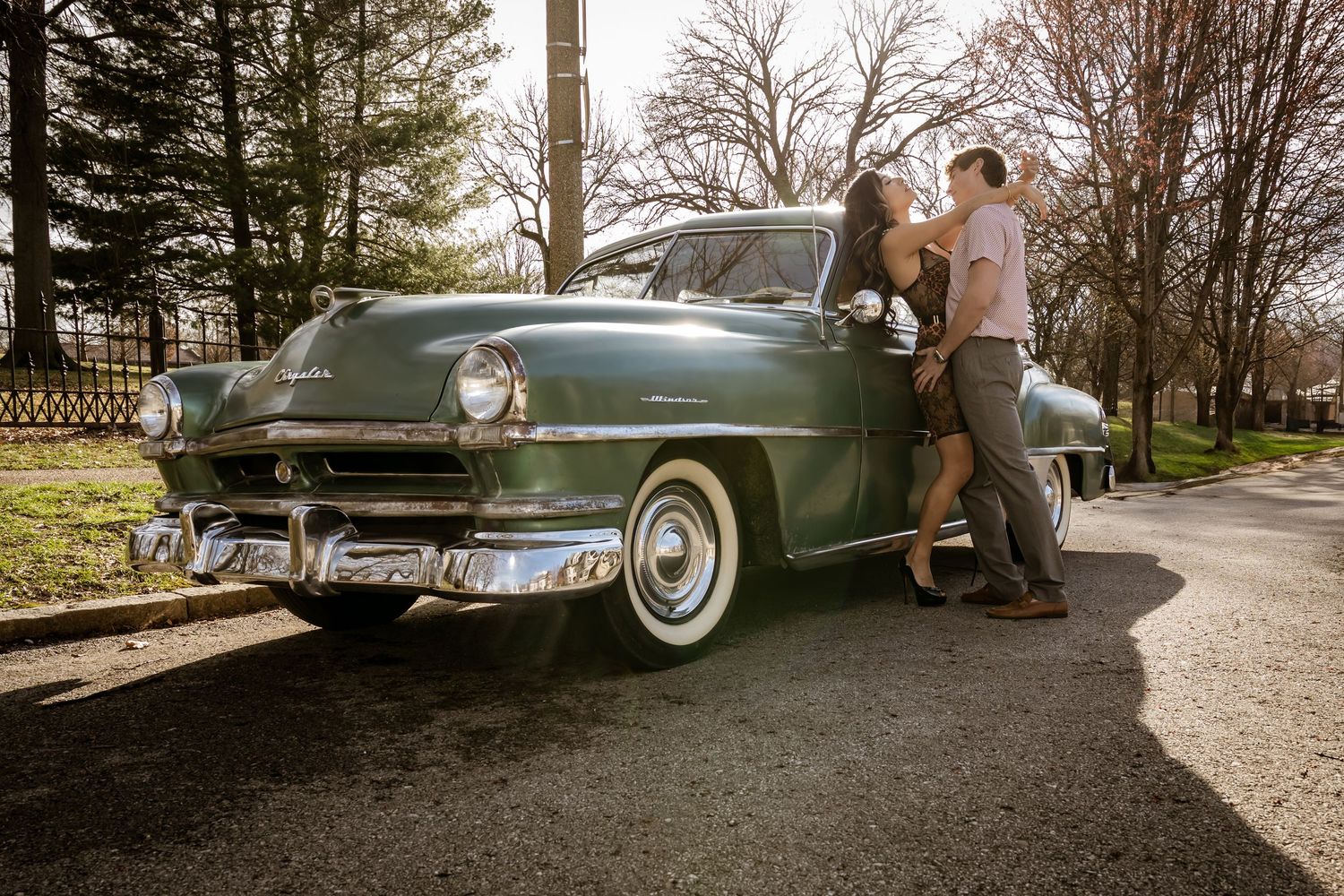Couple embracing beside a vintage teal classic car on a sunny day with bare trees in the background.