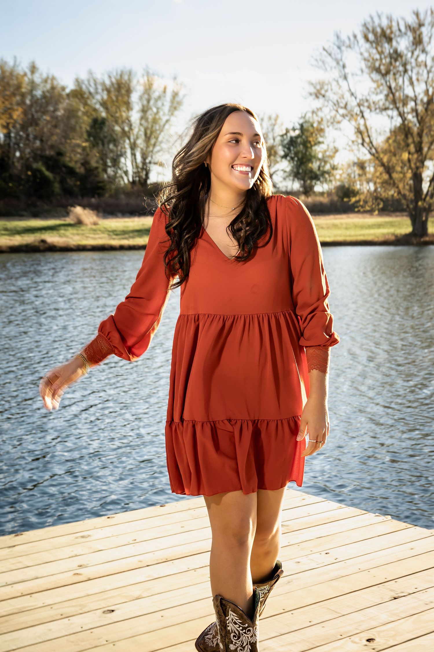 Woman in rust-colored blouse dancing joyfully on wooden dock by lake.
