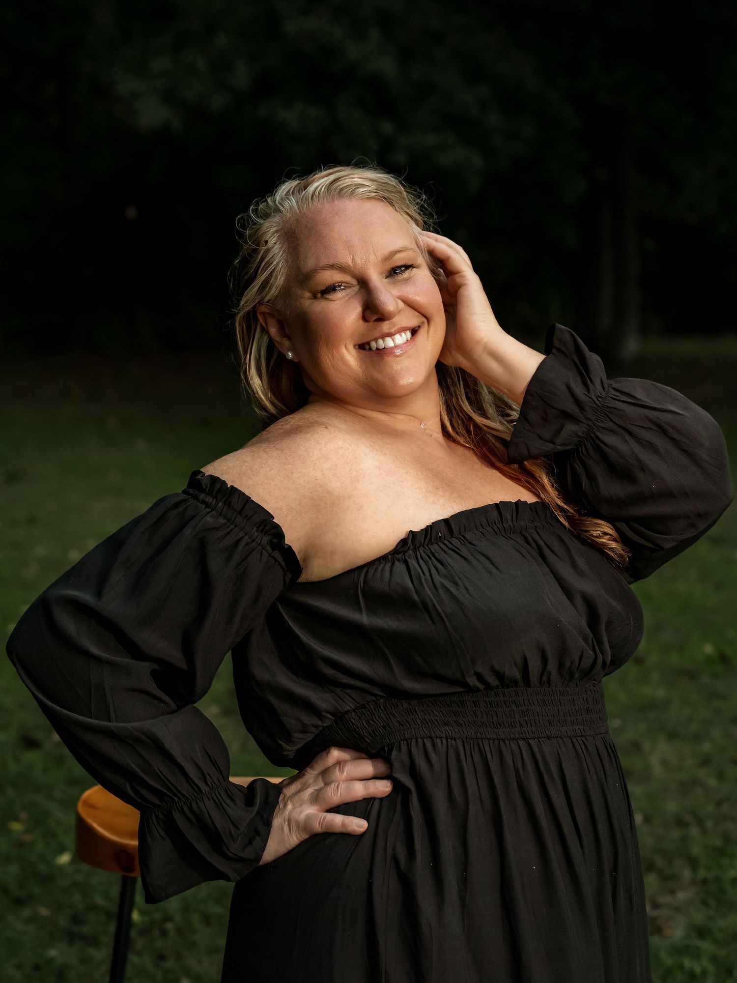 A smiling woman in a black off-shoulder dress poses outdoors with one hand on hip and other in hair.