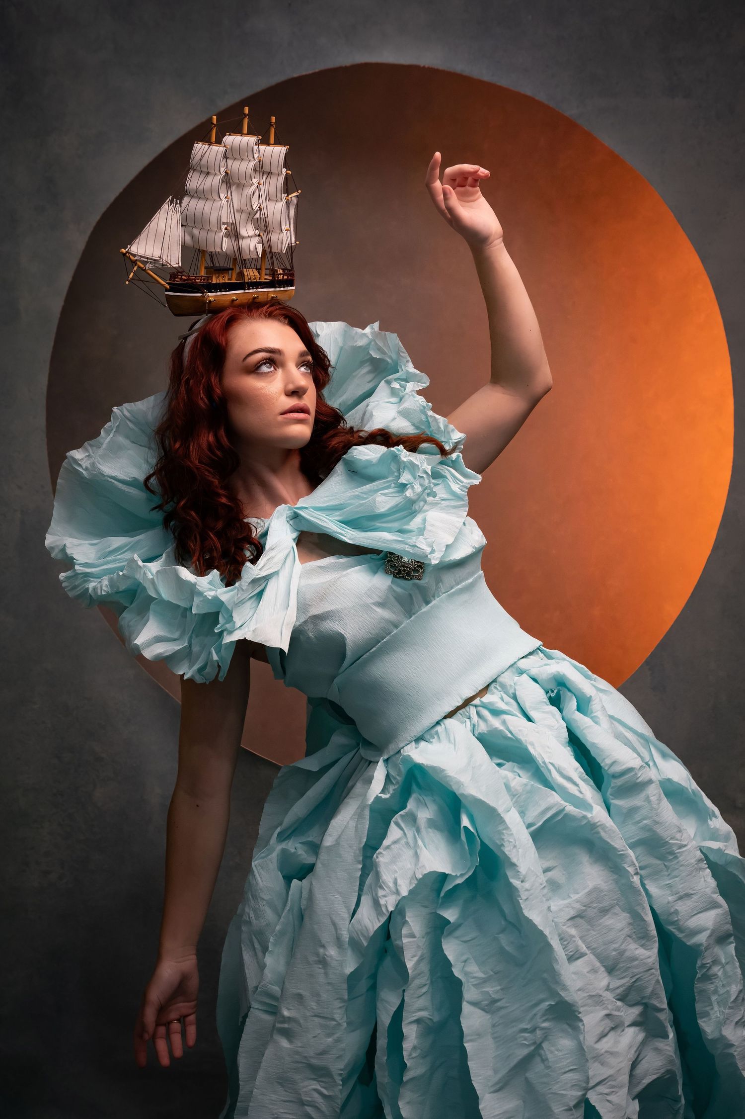 Whimsical portrait in light blue period dress with toy ship accessory against dark background.