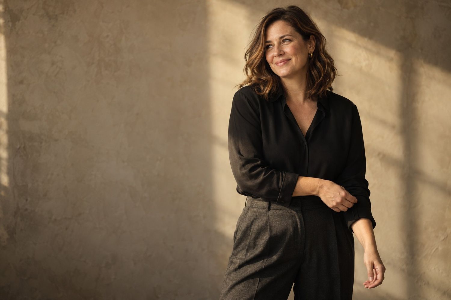 A smiling woman in a black blouse and gray trousers poses confidently against a warm beige textured wall.