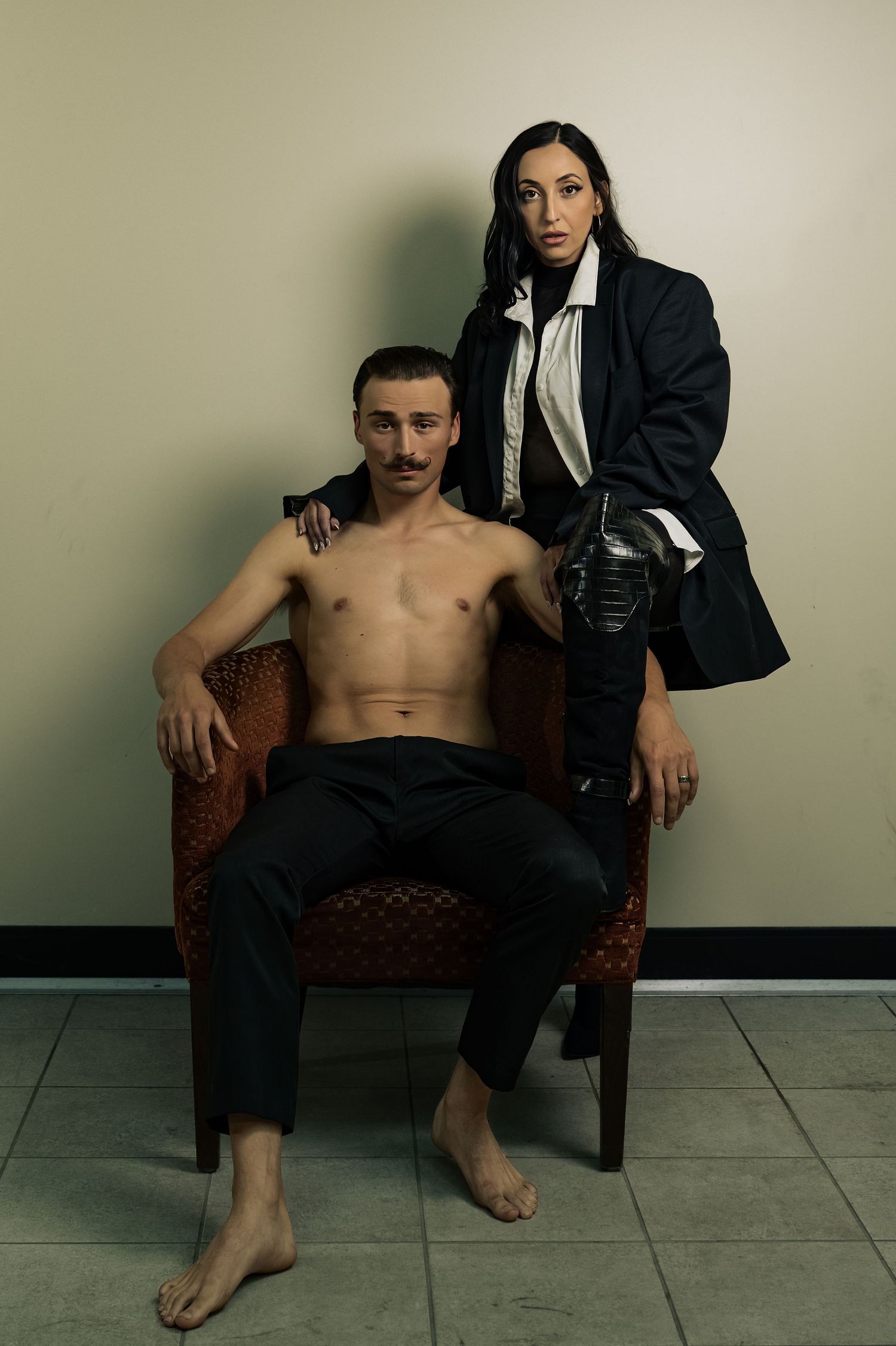 A shirtless man sits in a chair while a woman in a dark blazer stands behind him in a moody studio portrait.
