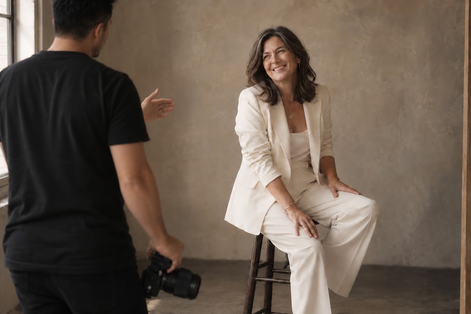 Photographer capturing a smiling woman in a white blazer and pants seated on a stool in a studio.