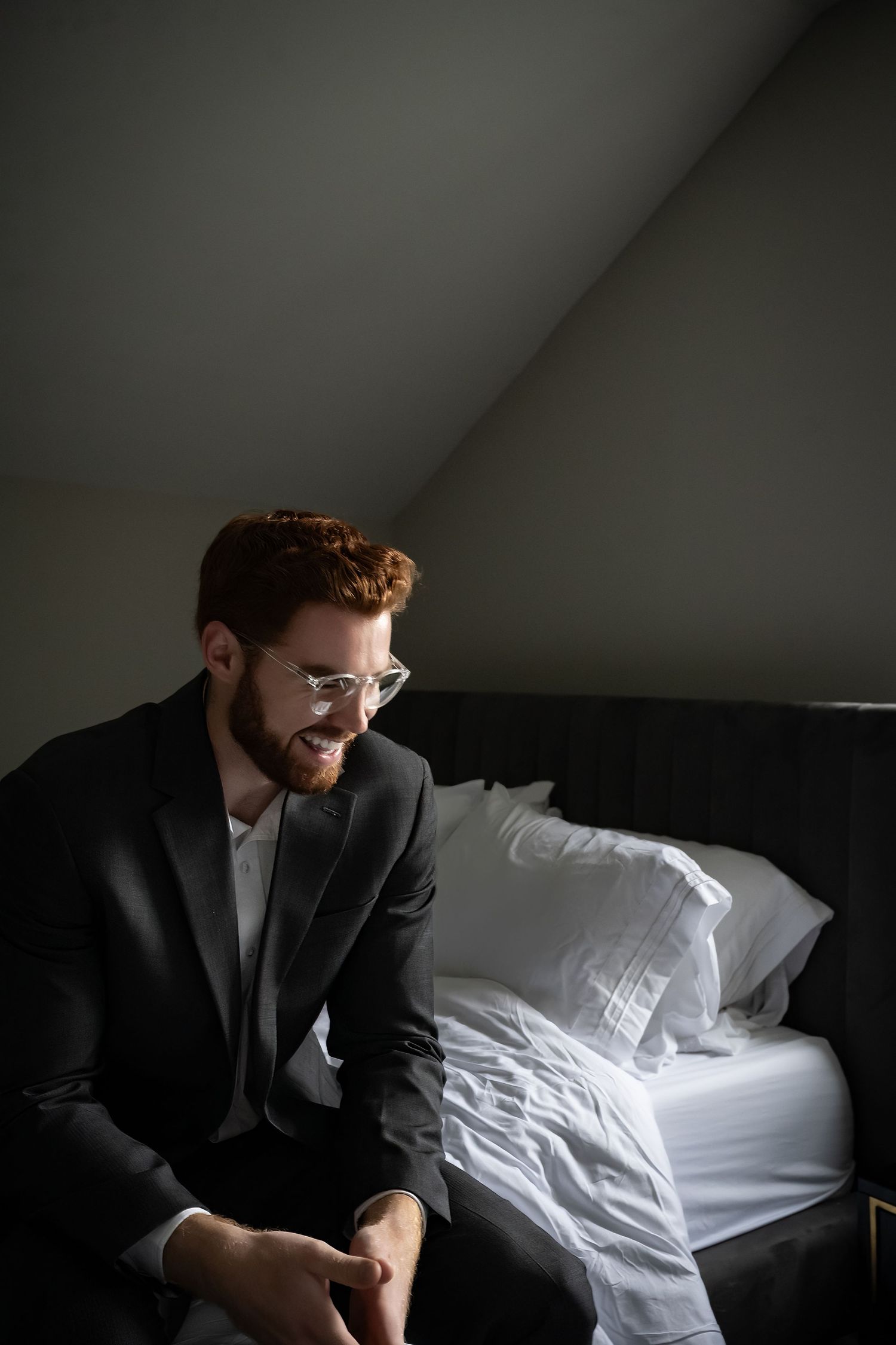 Bearded redhead man in black suit and glasses sitting on edge of bed in dark hotel room.