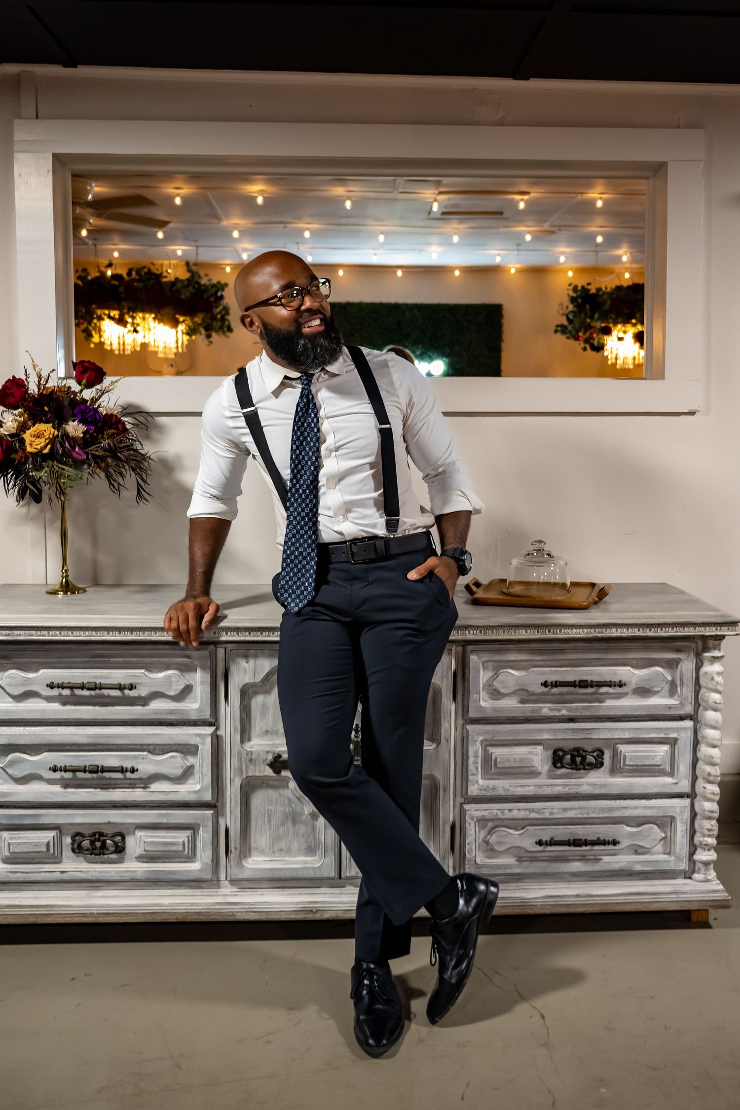 Smiling bald man in white shirt, suspenders, and tie leaning against vintage dresser with string lights backdrop.