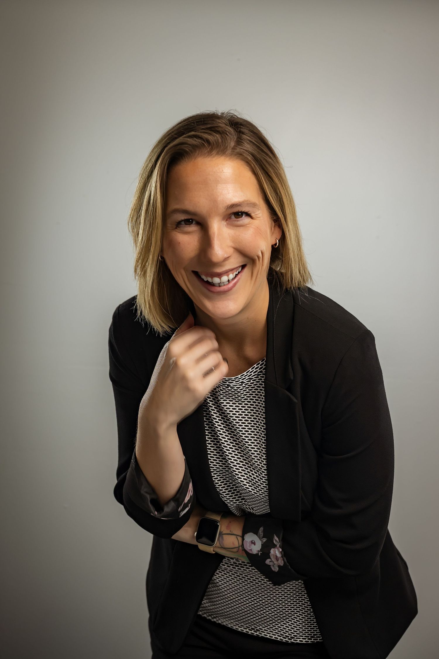 Smiling woman in black blazer and patterned top posing confidently against a light gray background.