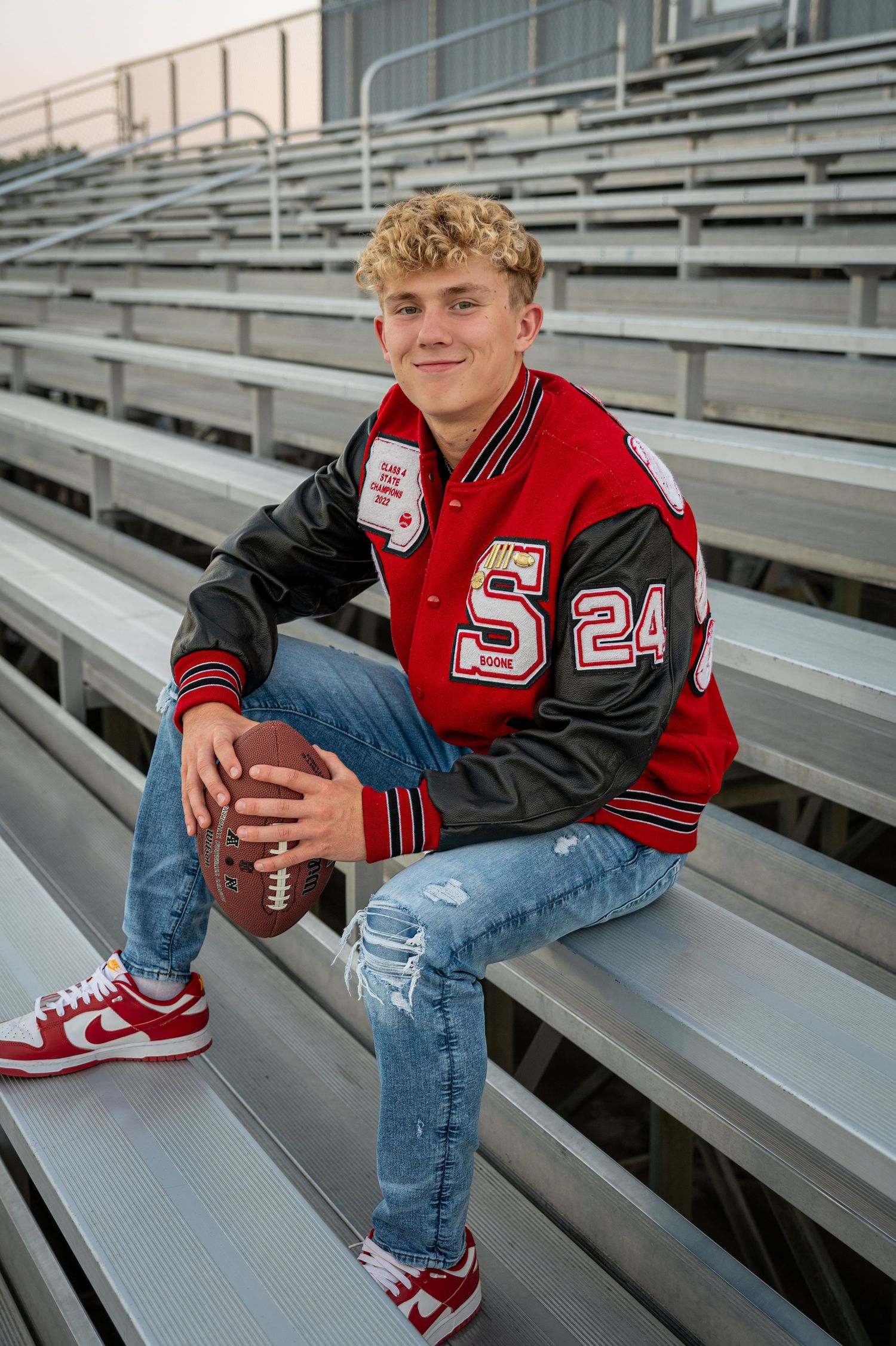 Athletic portrait on bleachers featuring varsity letterman jacket and football.