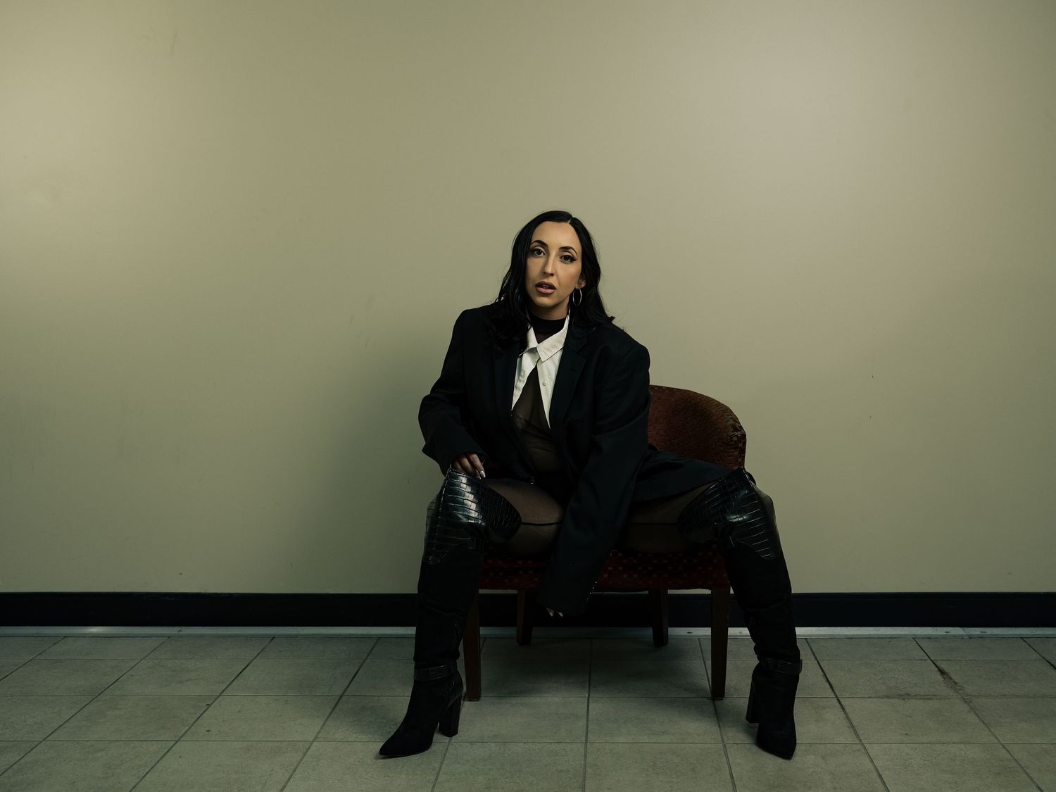 Woman in black blazer and boots sits confidently on a brown chair against a neutral wall.