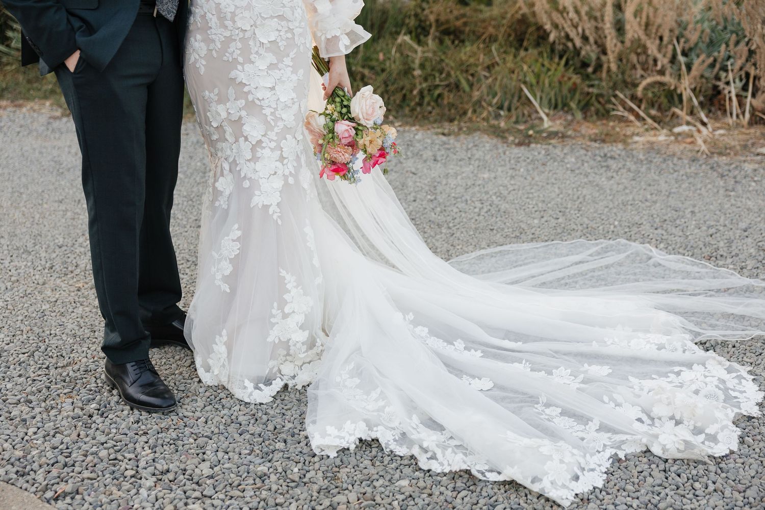 A detailed lace wedding dress train flows across gravel ground next to formal black shoes and a pink bouquet.