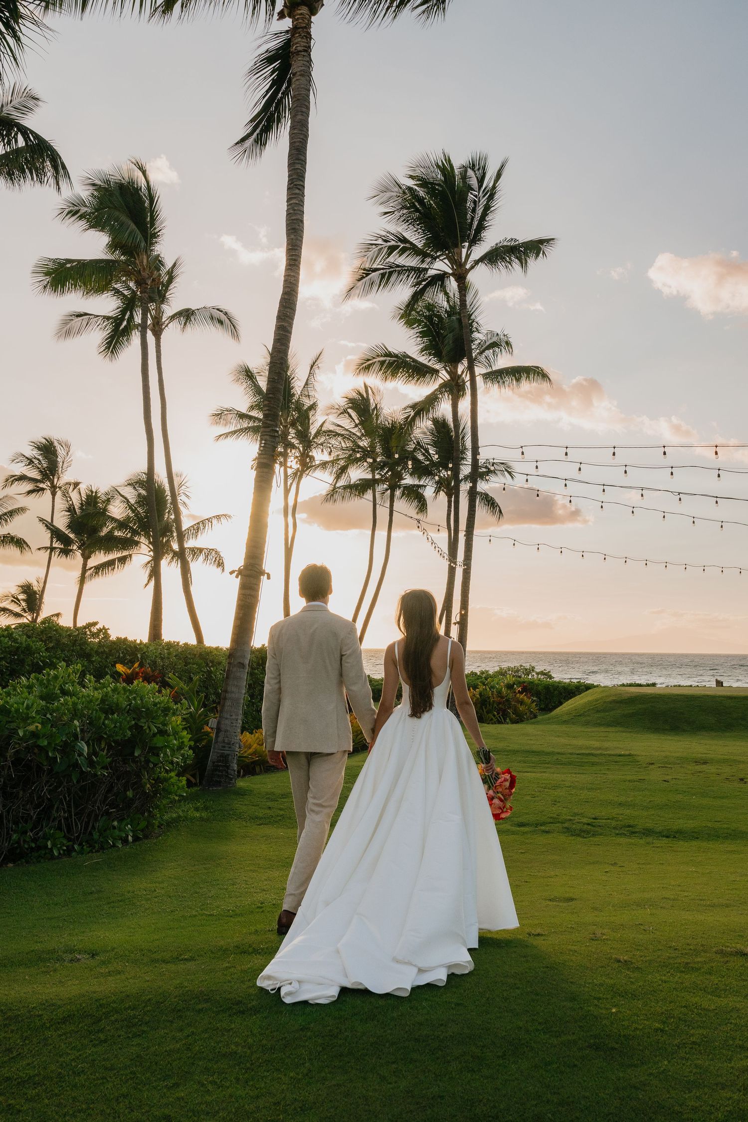 Wedding couple silhouetted against palm trees and string lights at a tropical beach during sunset.