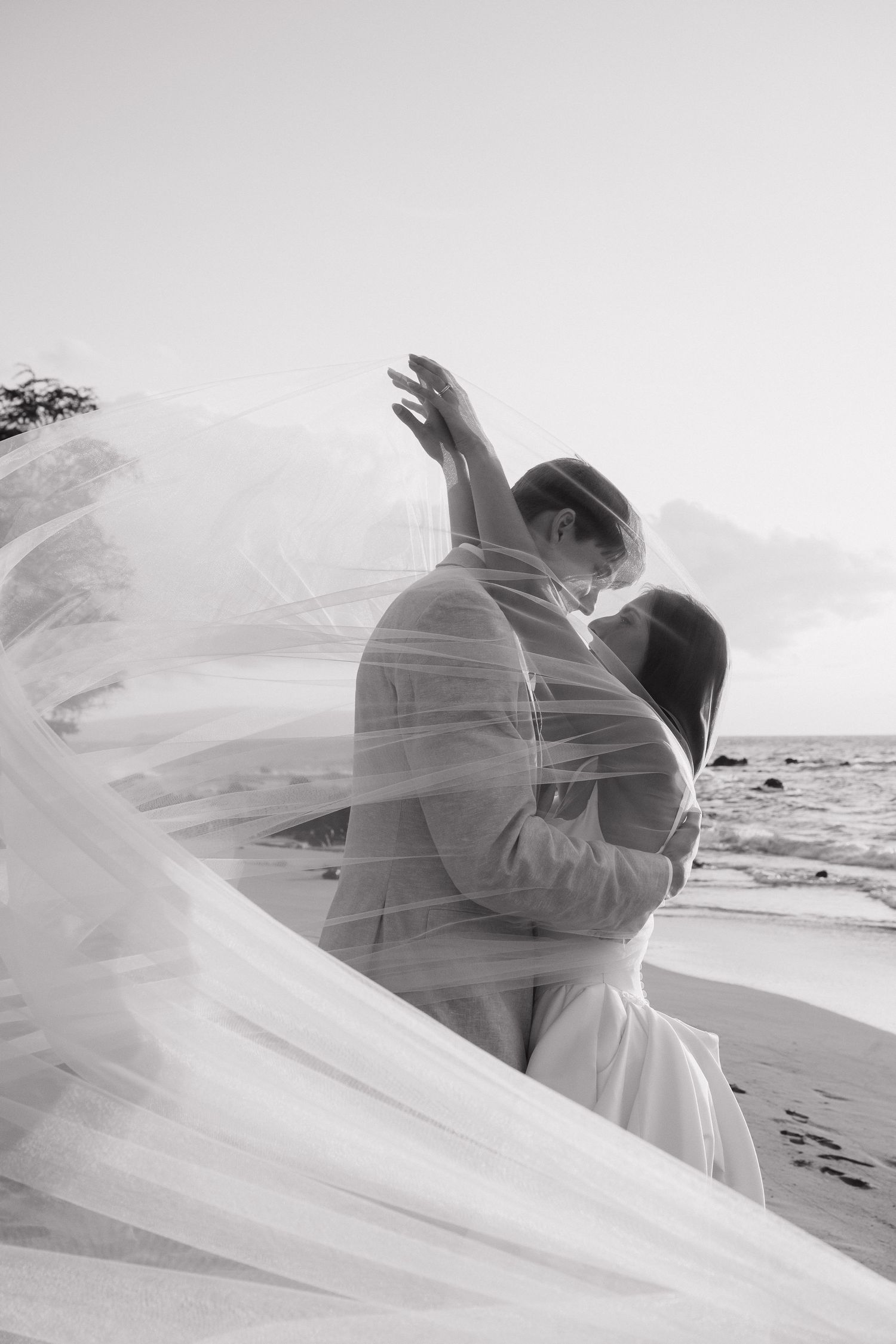 A couple embraces on a beach with flowing white veil creating an ethereal black and white silhouette against the ocean.