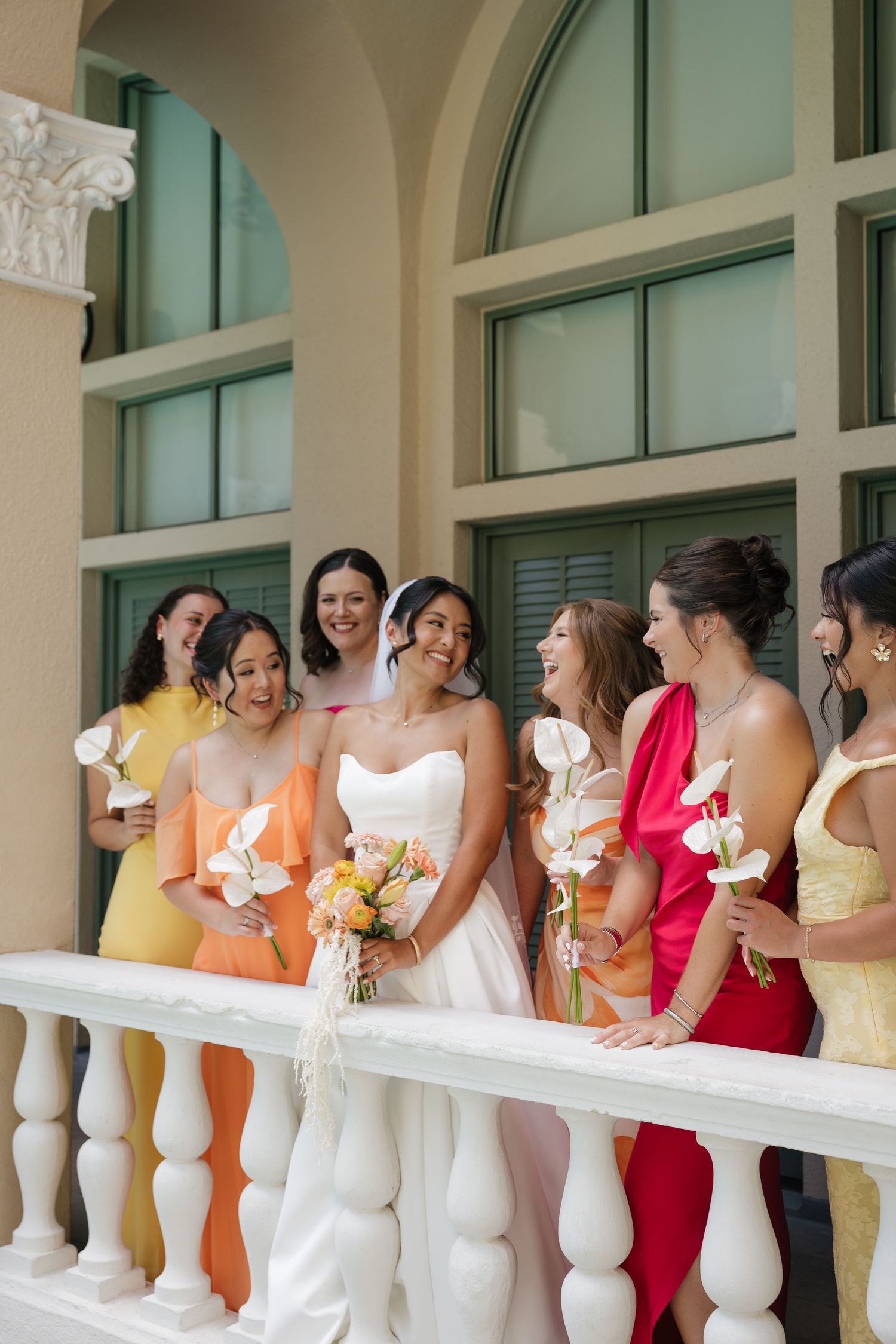 Bride in white strapless gown with bridesmaids in colorful mismatched dresses holding bouquets on elegant balcony.