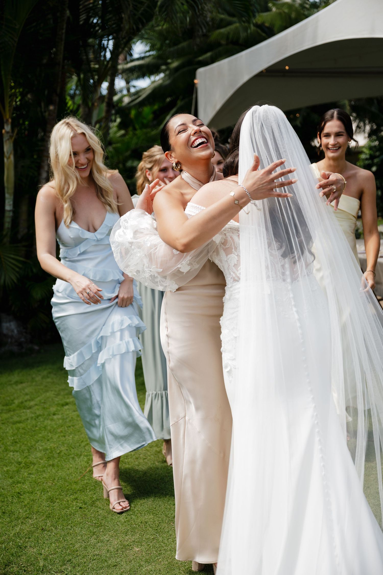 Wedding party celebrates joyfully on a grassy lawn with tropical palm trees in the background.
