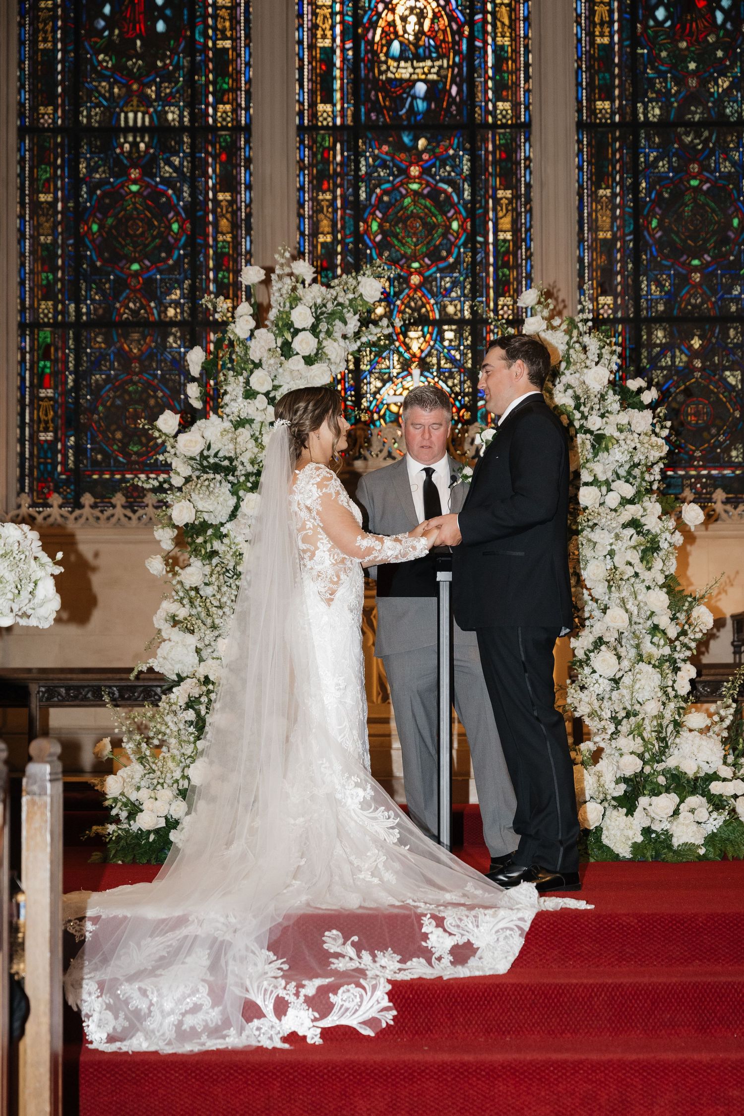 A wedding ceremony takes place in a church with stunning stained glass windows and white floral arrangements.