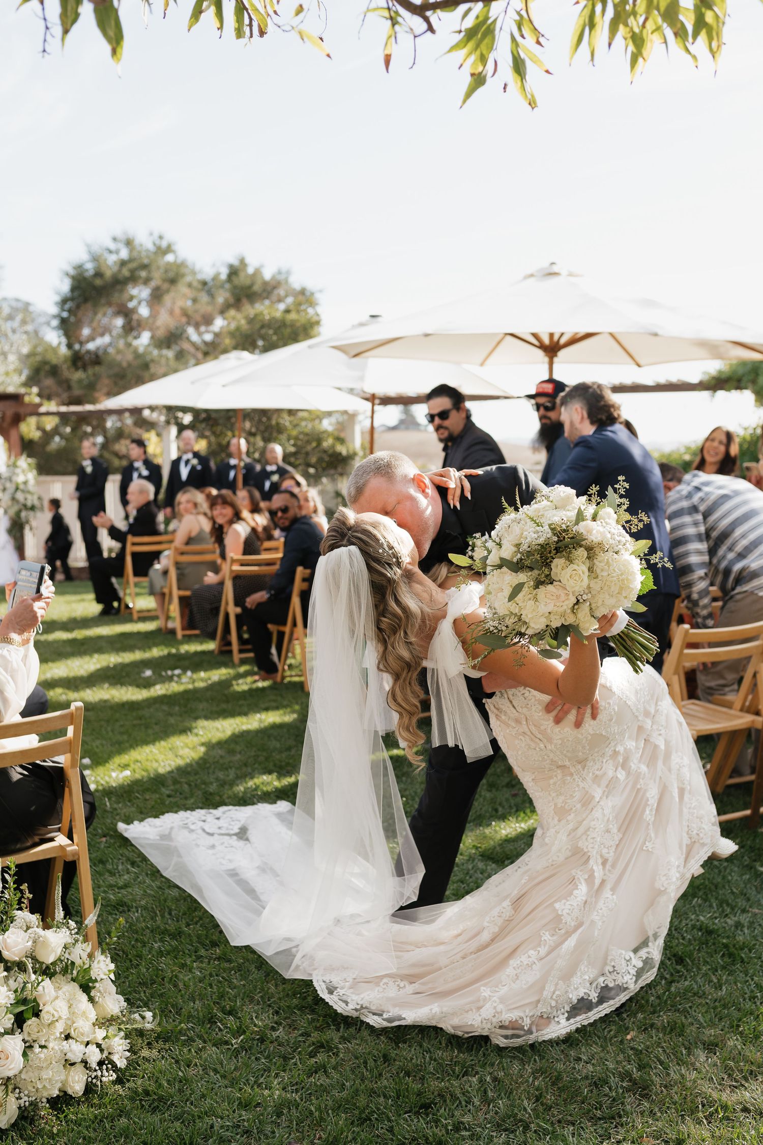 Romantic outdoor wedding ceremony moment with white floral arrangements and wooden chairs on grass lawn.
