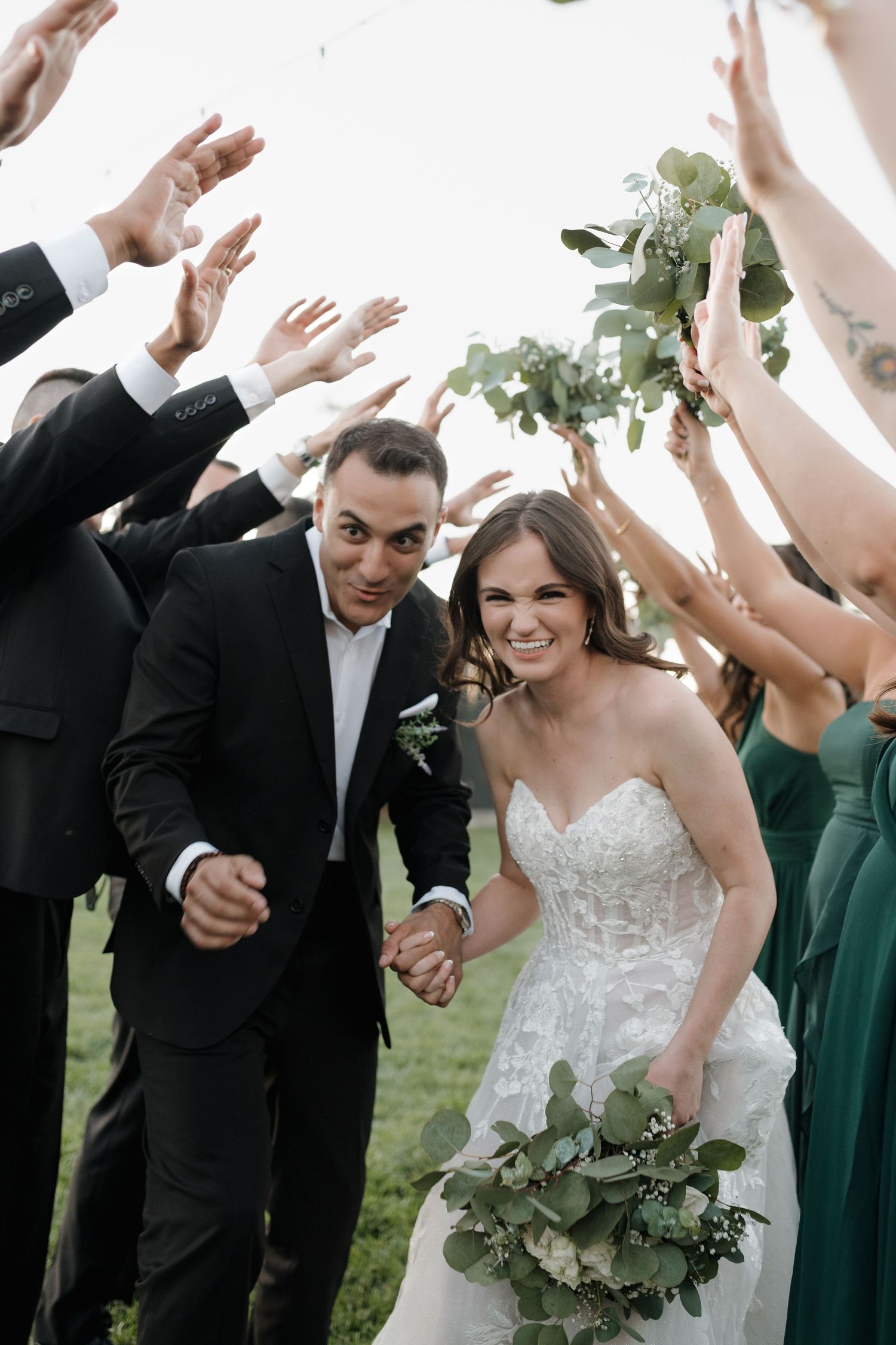Wedding party celebrates joyfully outdoors with raised arms as couple walks through in white dress and black suit.