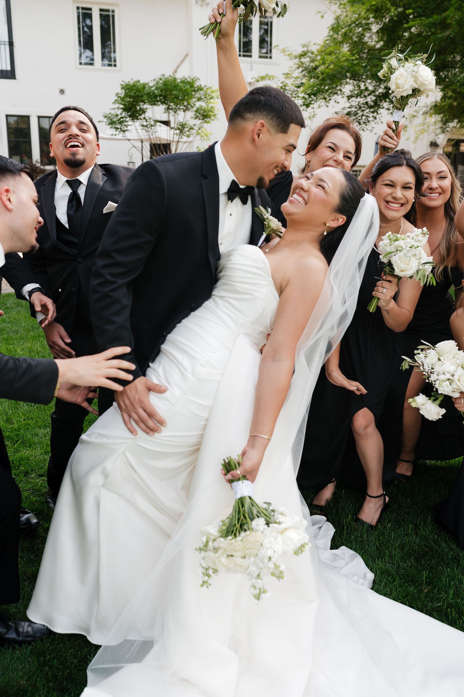 Joyful bride and groom share a romantic dip surrounded by smiling wedding party in black attire outdoors.