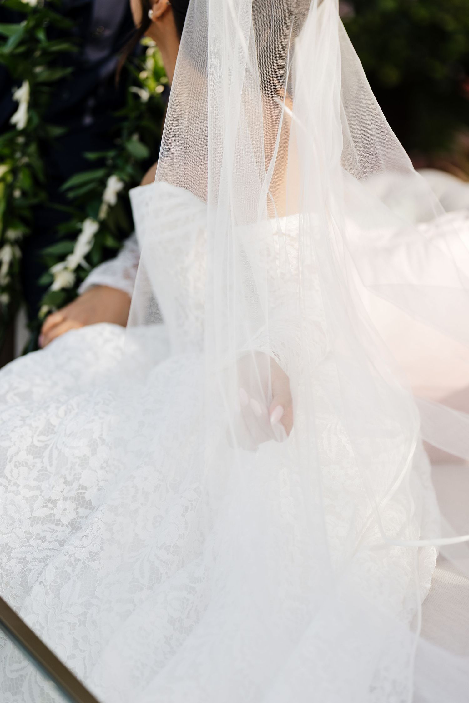 Close-up of a bride in a white lace wedding gown with a flowing veil at an outdoor ceremony.