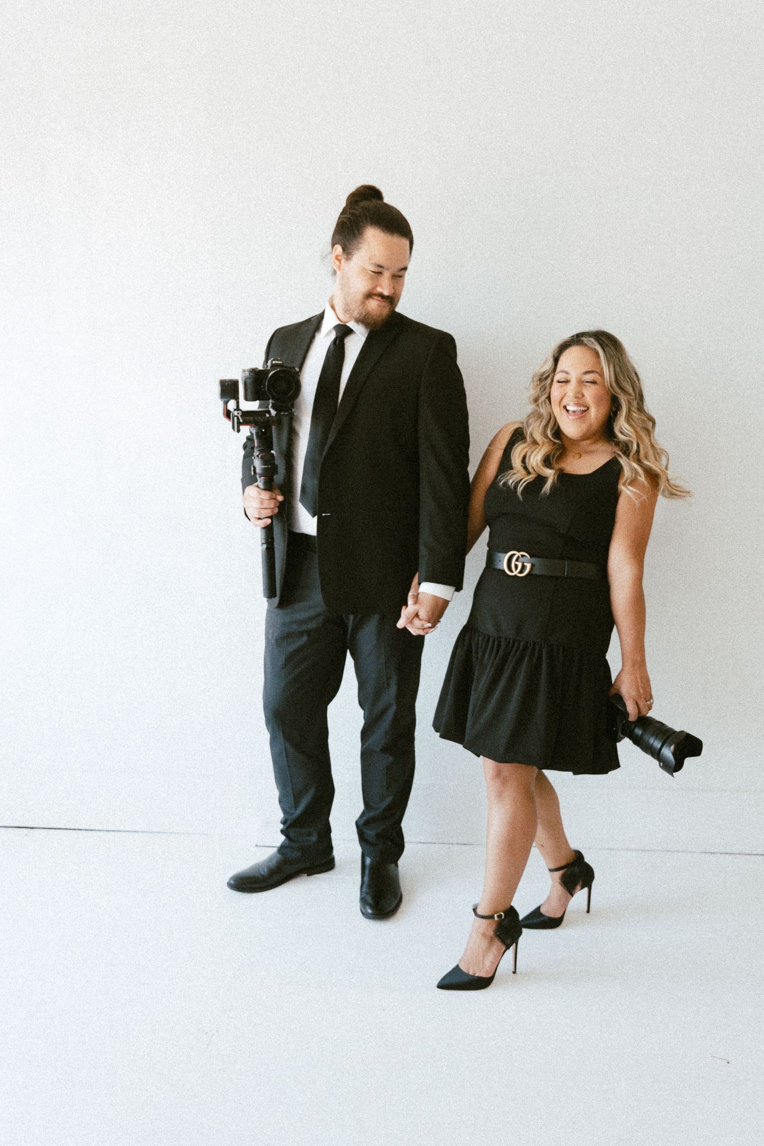 A professional photography duo poses playfully in black outfits with their camera gear against a white backdrop.