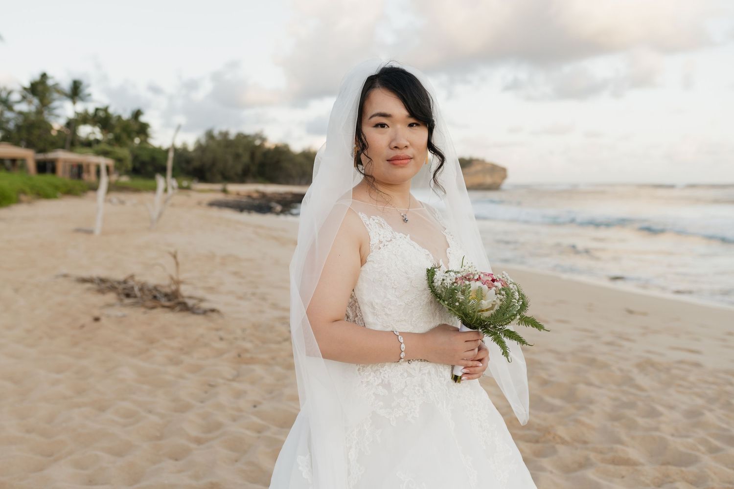 A bride in a white dress holding a bouquet on a sandy beach at sunset.