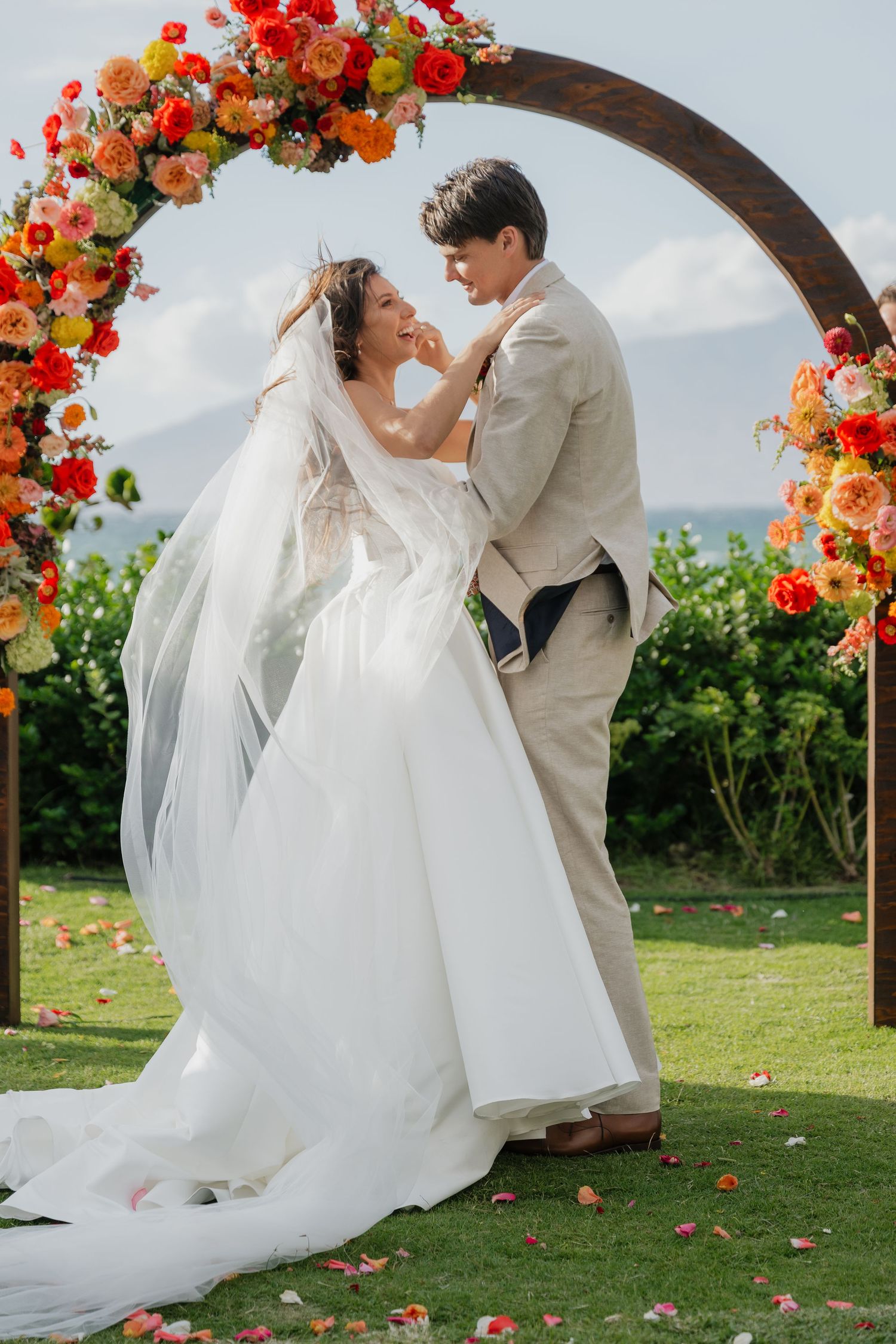 A couple shares a romantic moment under a floral arch with vibrant red and orange blooms.