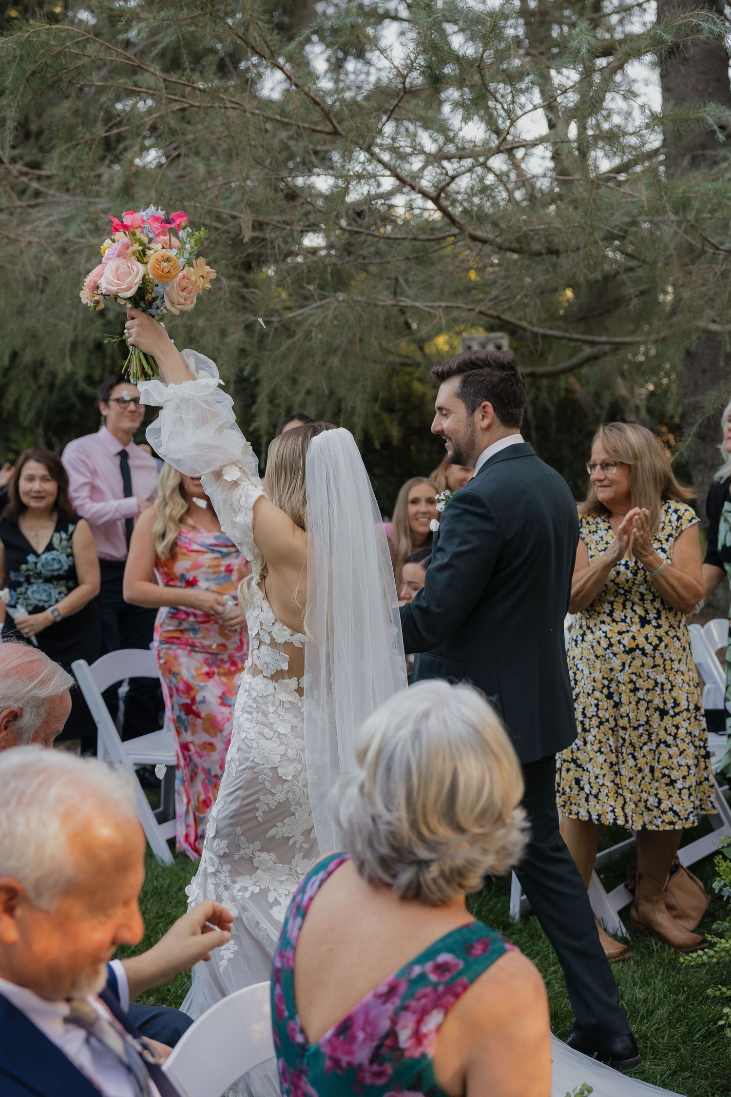 An outdoor wedding ceremony takes place under tall evergreen trees with guests seated on white chairs watching the couple exchange vows.