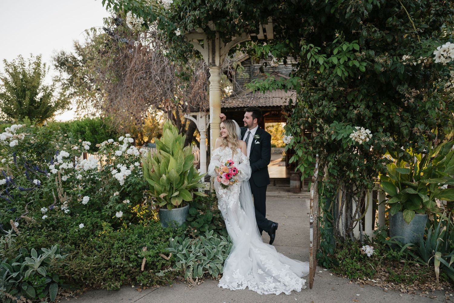 A couple poses in a lush garden setting with romantic lighting and floral decor at an outdoor wedding venue.