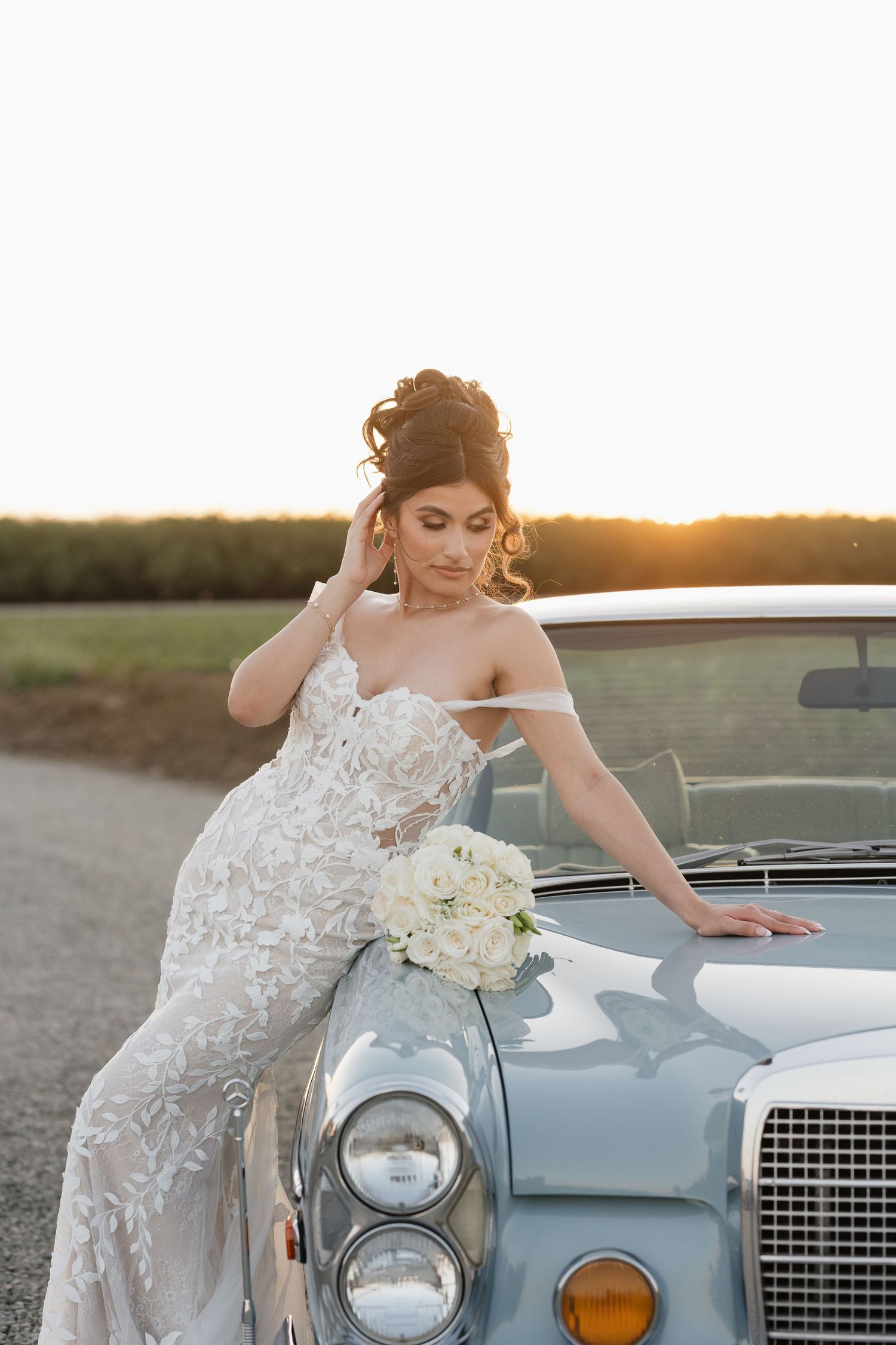 A bride in a lace gown poses against a classic car during golden hour.