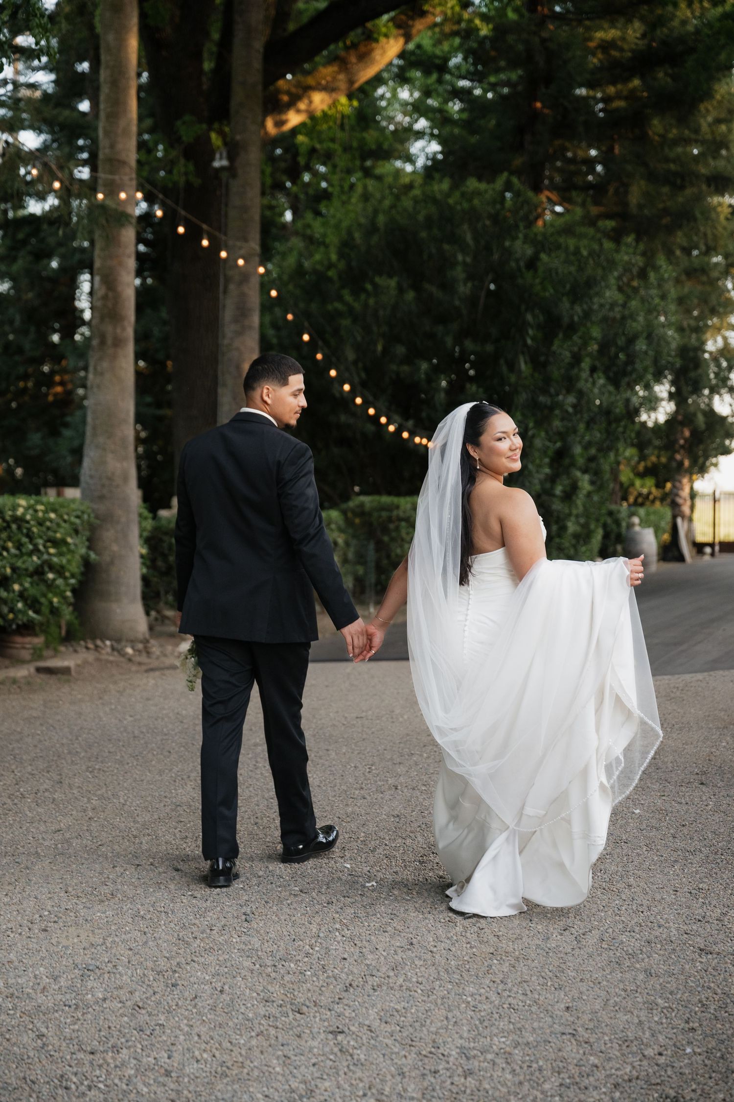 Bride and groom walk hand-in-hand outdoors, bride in white gown with veil, groom in black suit, string lights above.