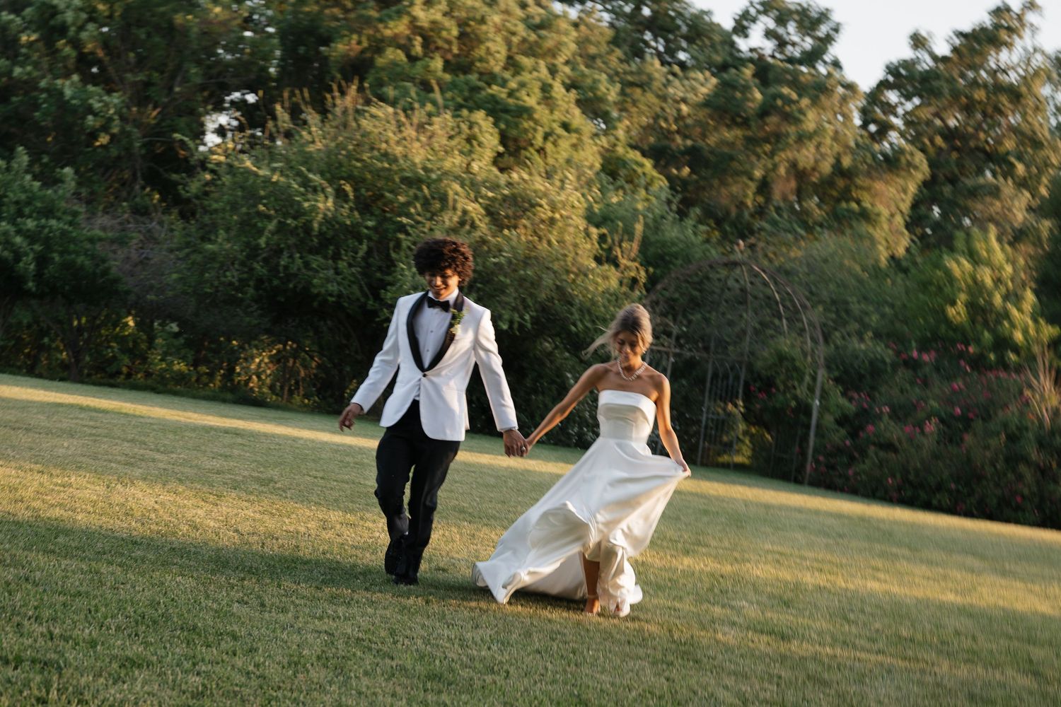 Newlyweds walk hand in hand across sun-drenched field during golden hour.