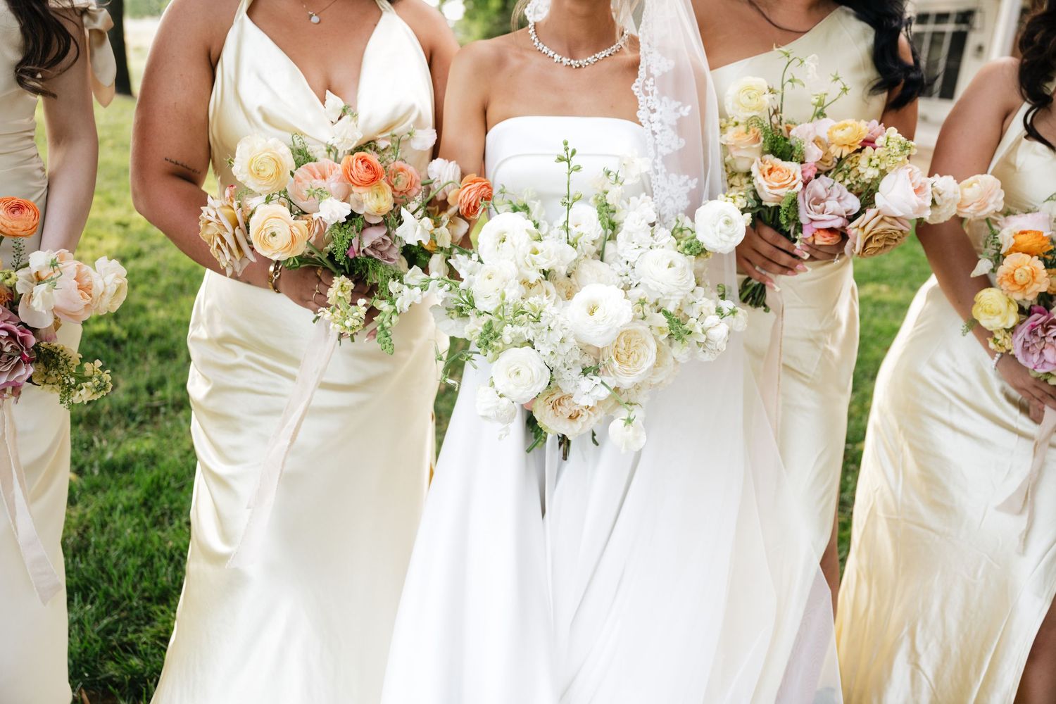 Classic white-gowned bridesmaids hold elegant cream and white floral bouquets in an outdoor wedding setting.