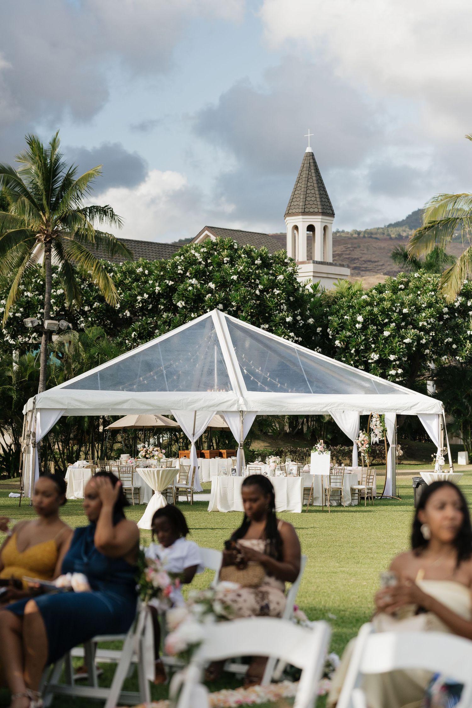 Wedding guests enjoy an outdoor reception under white tents with palm trees and mountains in the background.