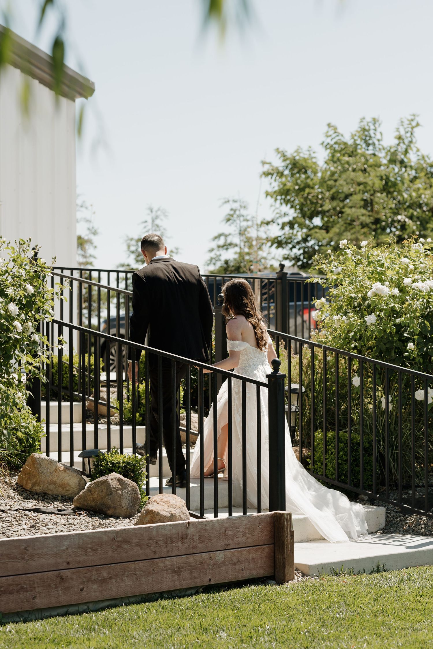 A wedding couple walks along a landscaped path with black railings and flowering bushes.