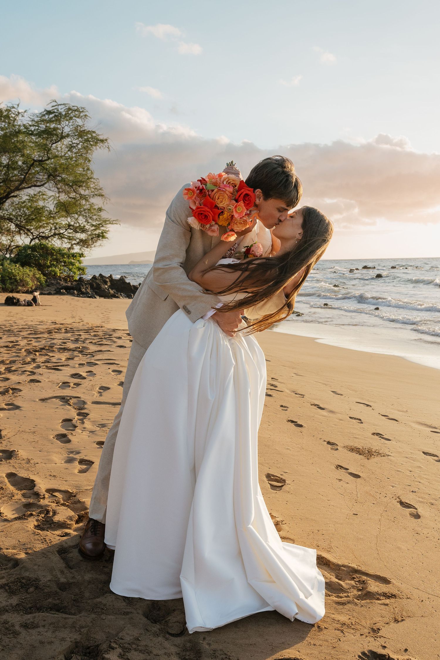 A couple dips and kisses on a sandy beach at sunset with tropical trees in the background.