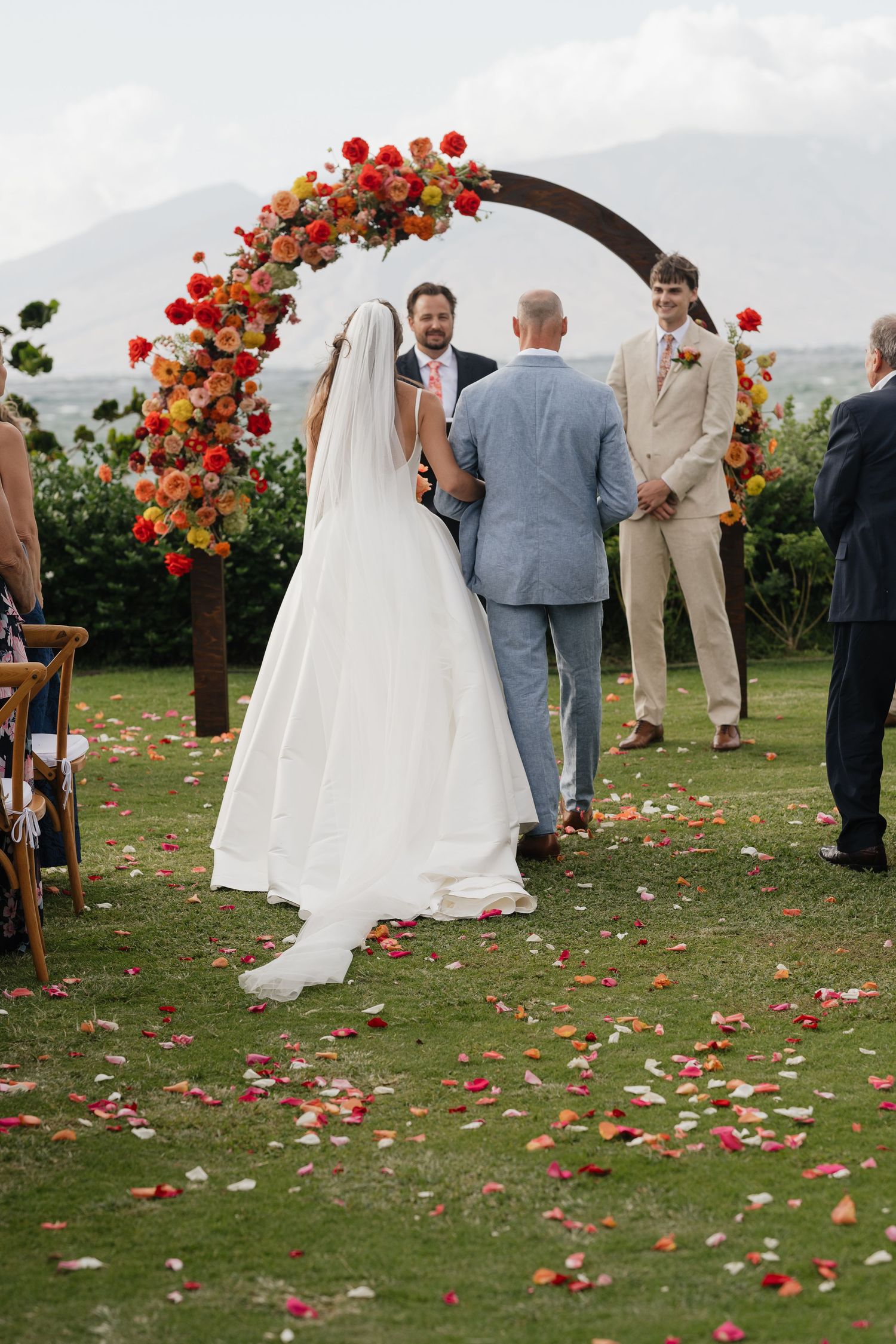 Wedding ceremony setup with red floral arch and scattered petals on grass with mountain views.