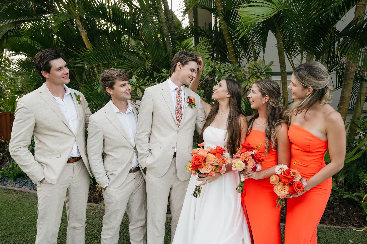 Wedding party in beige suits and coral bridesmaid dresses gather outdoors in a tropical garden setting.