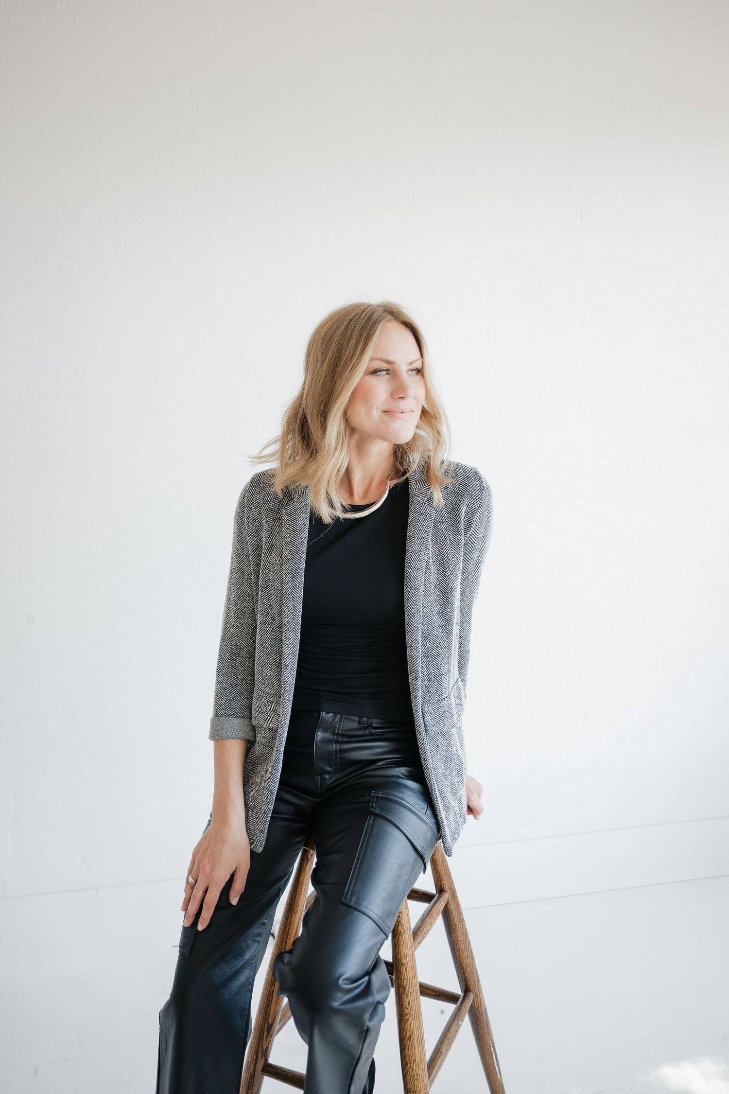 Professional fashion portrait of someone seated on wooden chair wearing gray blazer and black outfit against white background.