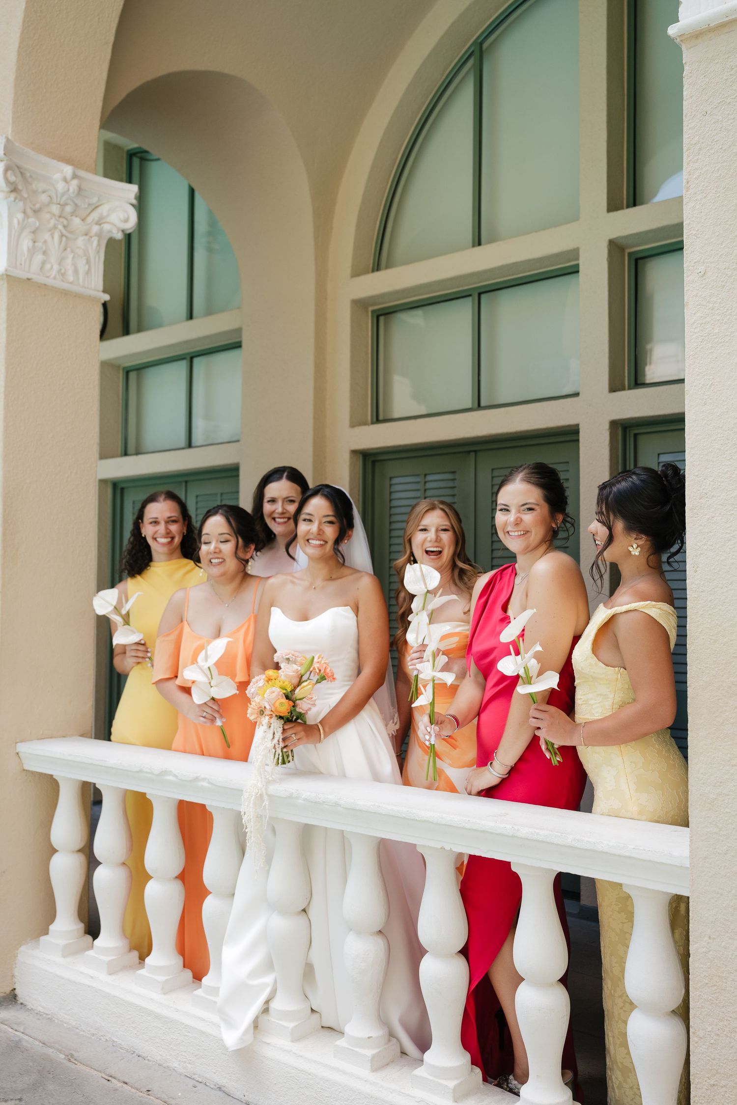 Bride in white gown with bridesmaids in colorful yellow, orange, and red dresses holding calla lilies on elegant balcony.