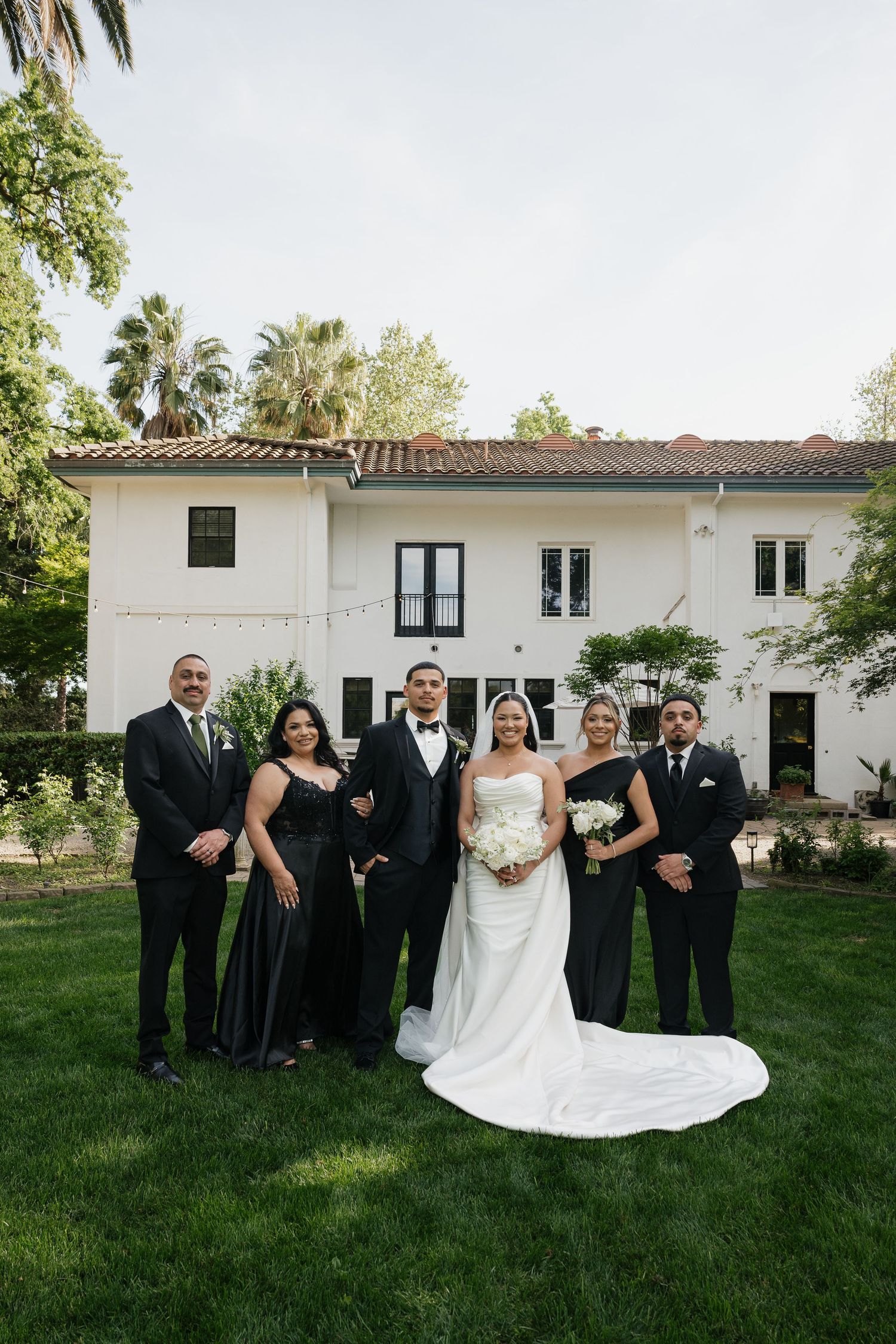 Wedding party poses in black attire with bride in white gown before elegant white Spanish-style estate.